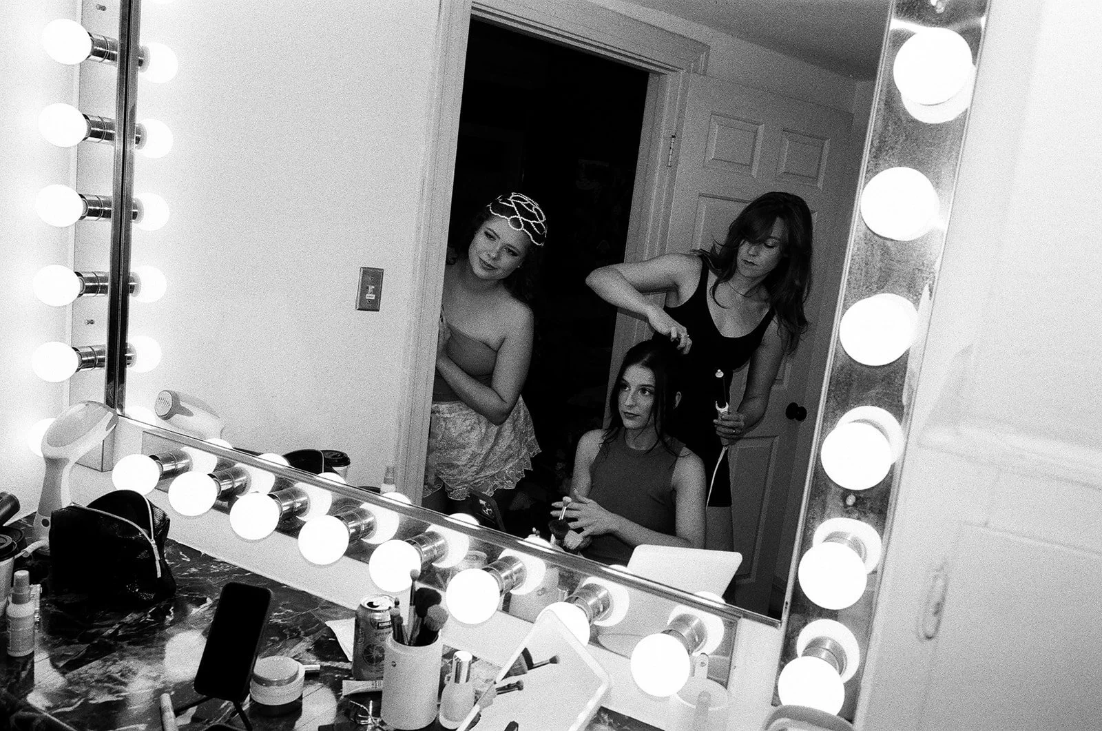 Two women backstage, one sitting getting her hair styled and the other standing holding a styling tool, visible in a makeup mirror surrounded by lights.