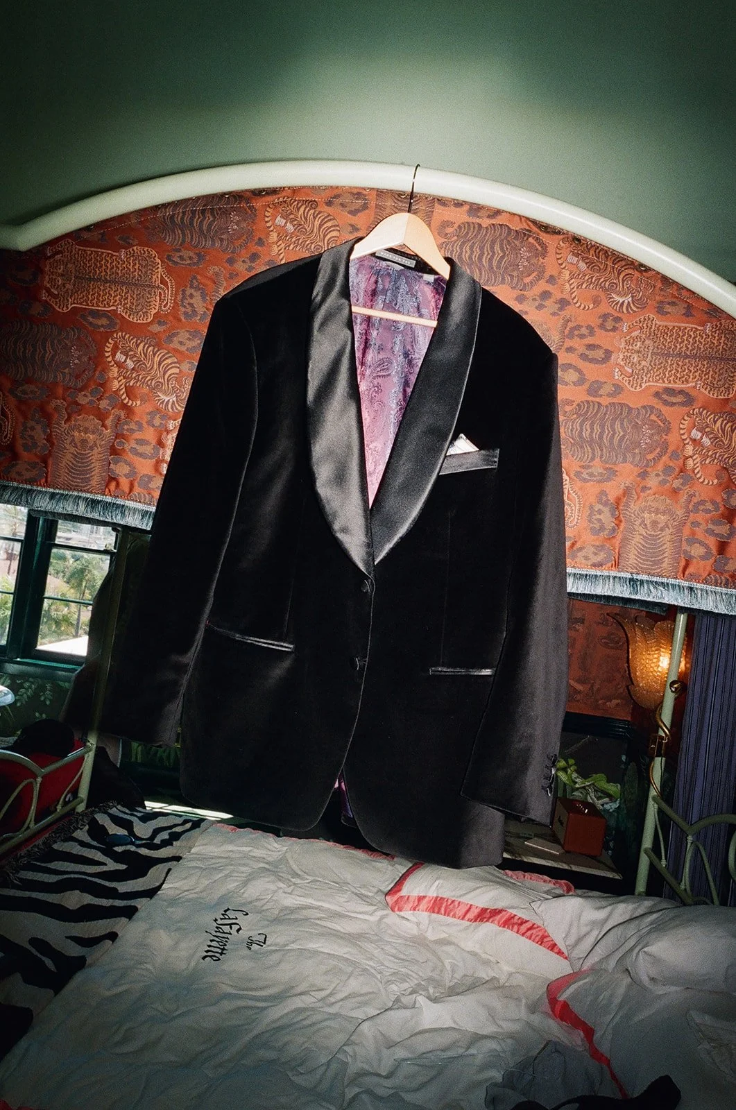 A black tuxedo jacket hanging on a wooden hanger from a curved headboard in a bedroom with patterned wallpaper and bed linens.