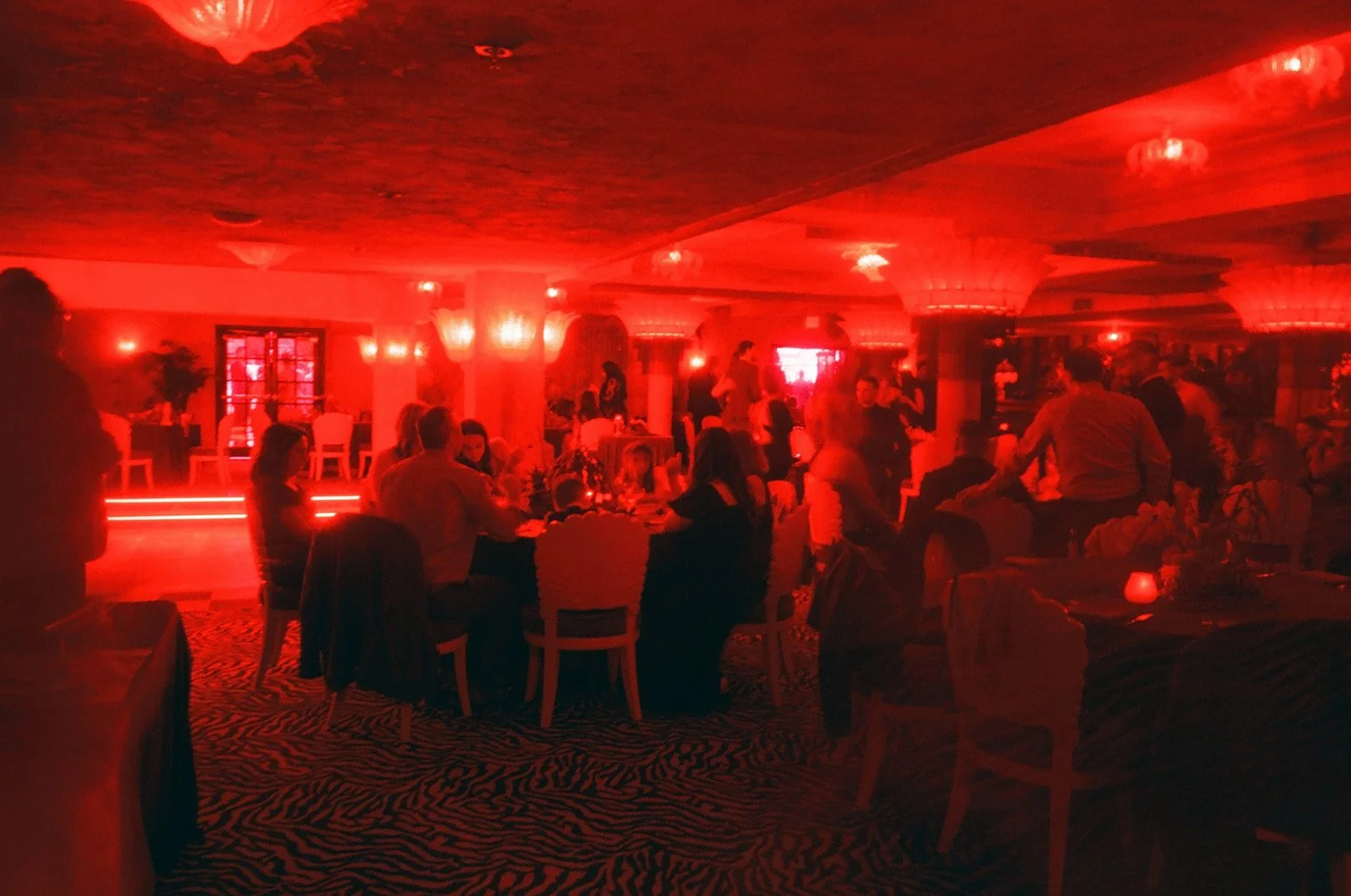 Dimly lit restaurant or bar with red lighting, people sitting at tables, some standing and socializing, decorative zebra print carpet, candles on tables.