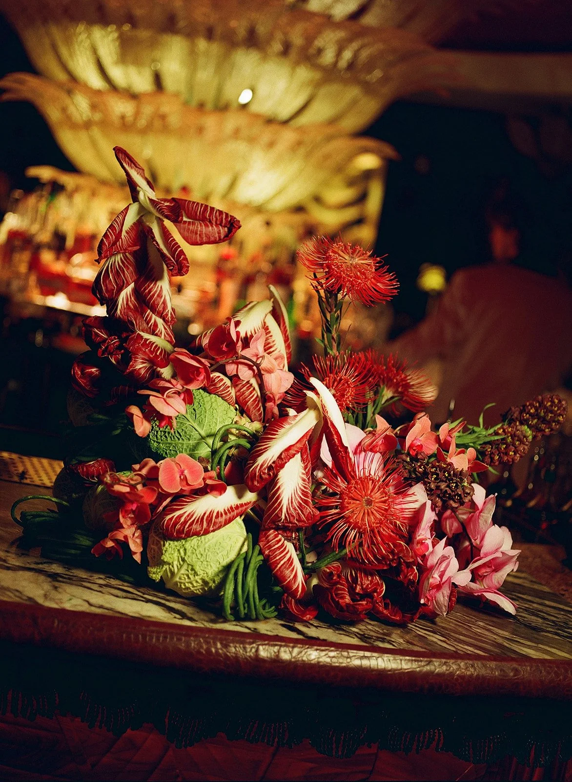 A vibrant floral arrangement featuring red and pink flowers on a decorative wooden surface with a blurred ornate background.
