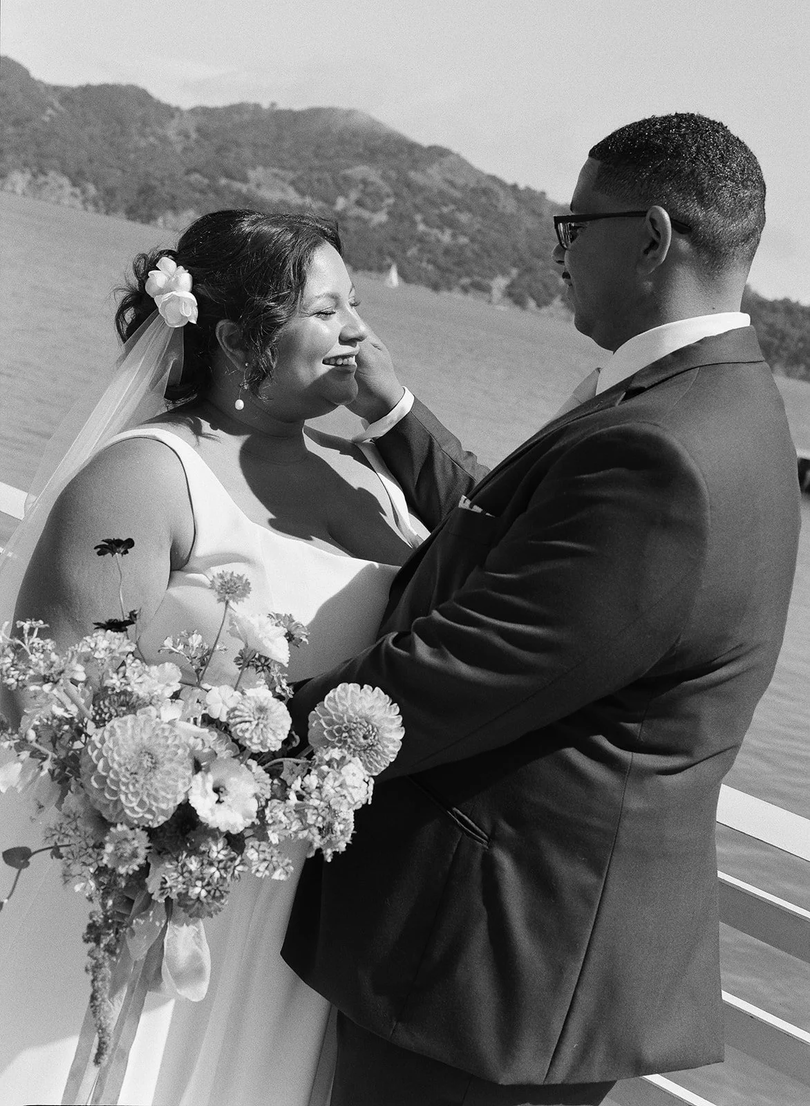 A joyful bride and groom on their wedding day, embracing outdoors near water with mountains in the background, the bride holding a bouquet of flowers.