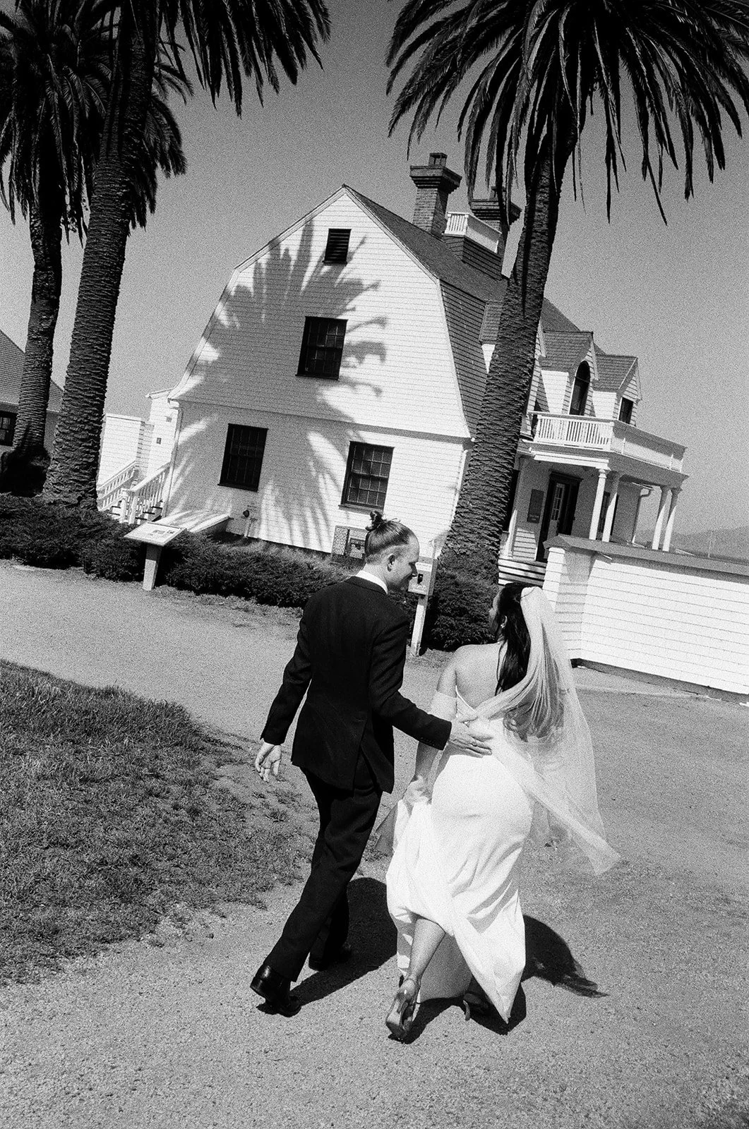A black-and-white photo of a bride and groom walking hand-in-hand outside a house with palm trees.