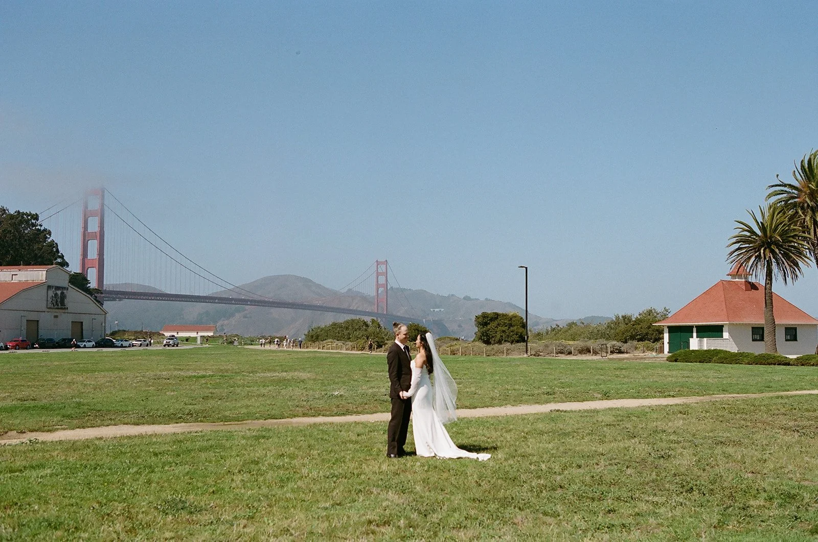 A bride and groom holding hands and facing each other in a green field with the Golden Gate Bridge in the background on a clear day.