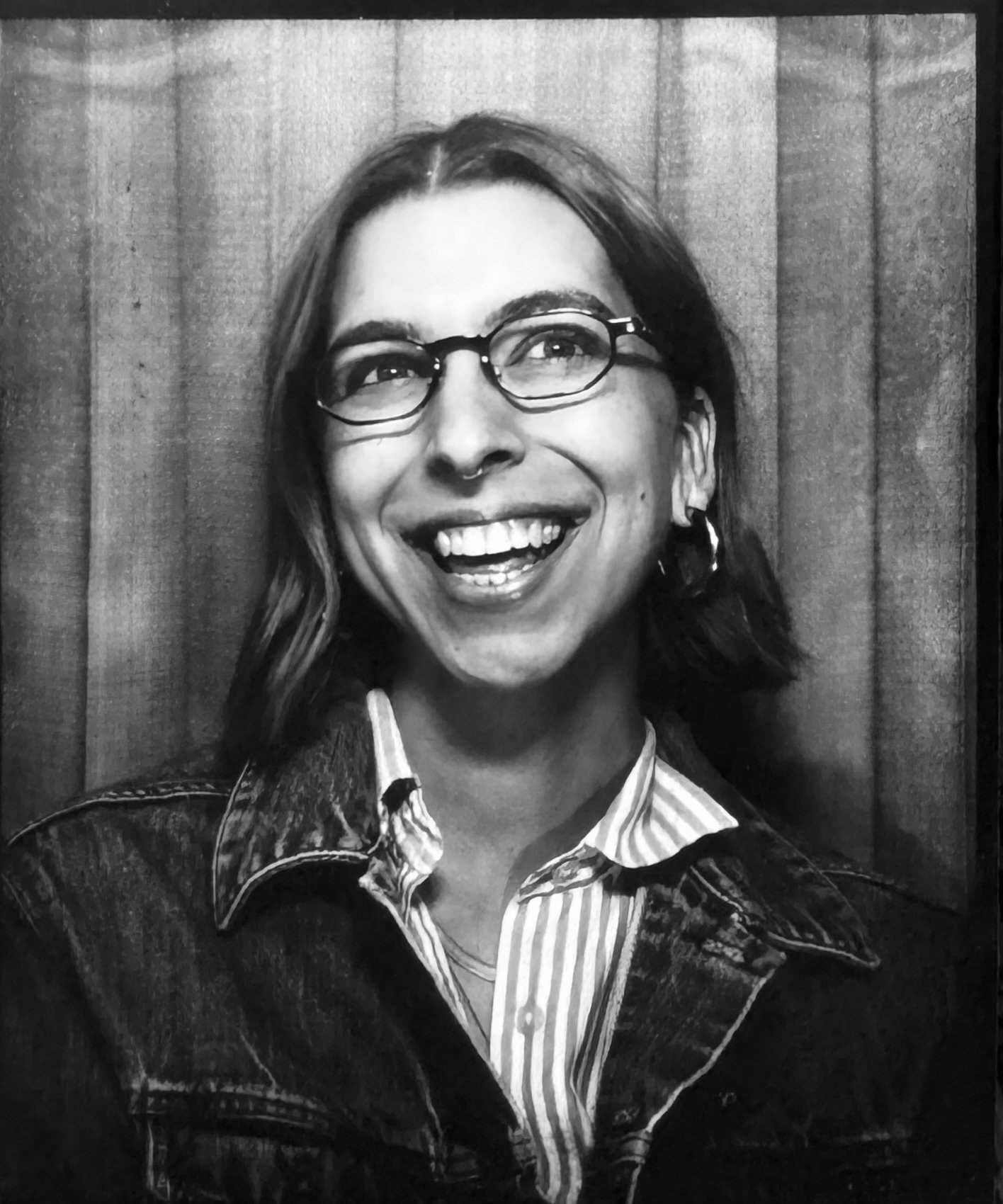 A smiling woman with glasses, earrings, and shoulder-length hair, wearing a striped shirt and denim jacket, posed against a wooden background.