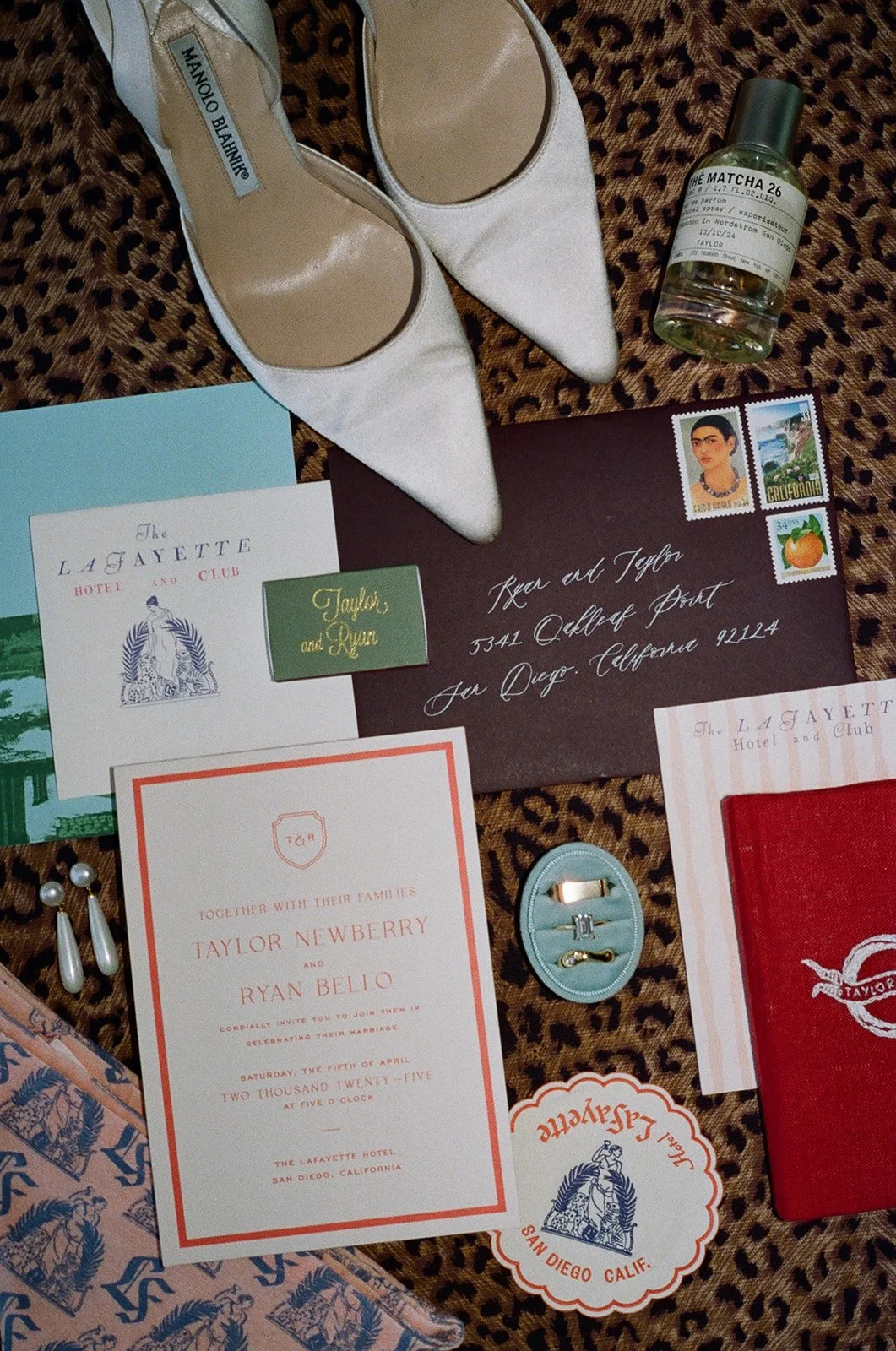 Flat lay of wedding or event stationery, shoes, jewelry, and perfume on a leopard print surface. Includes a white pair of satin pointed shoes, a green box of rings, a bottle of perfume, a wedding invitation, an envelope, and various cards and tickets