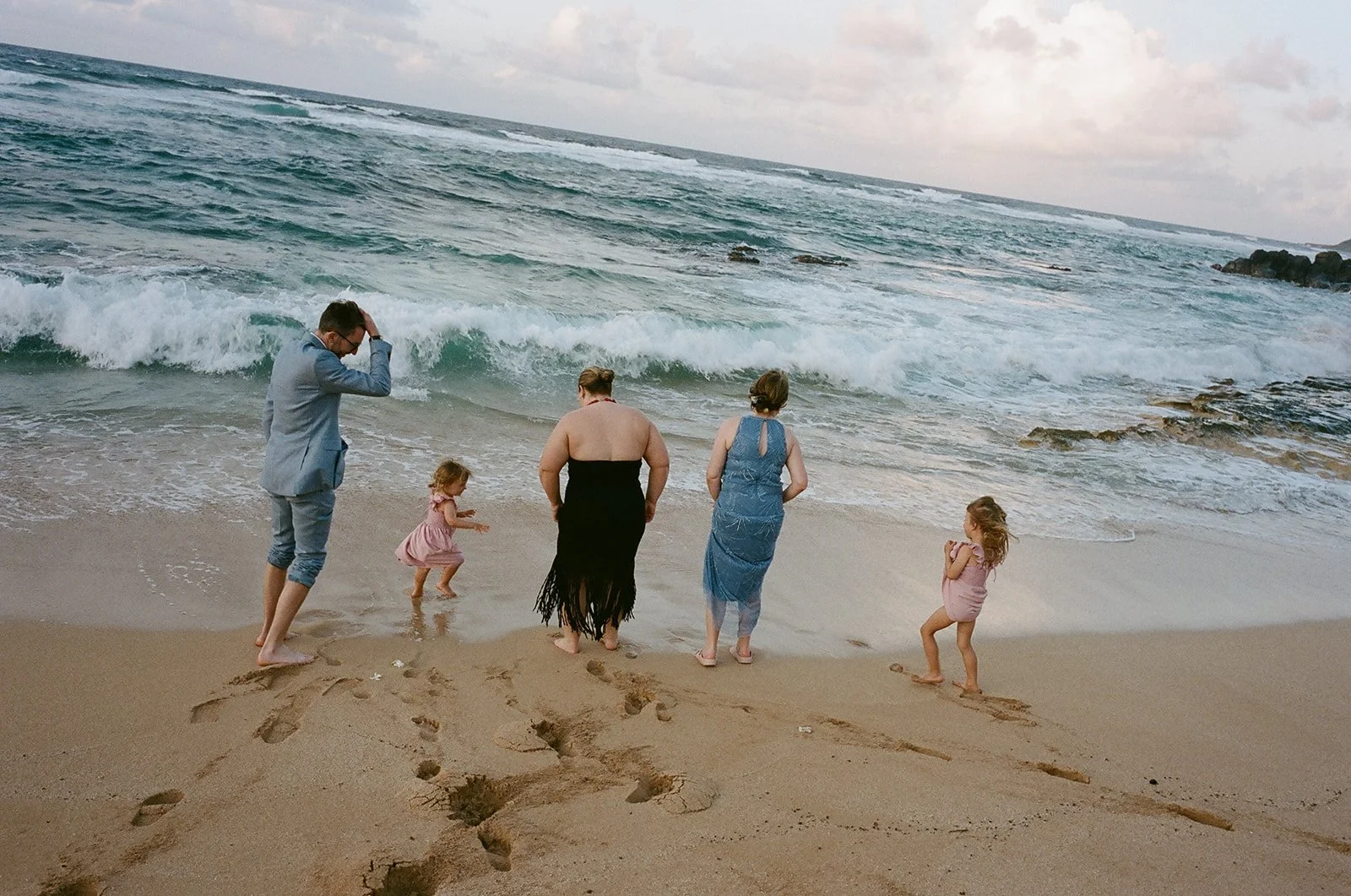 Family of five on the beach, two young girls, two women, and a man, standing near the water's edge with waves crashing, footprints in the sand, overcast sky.