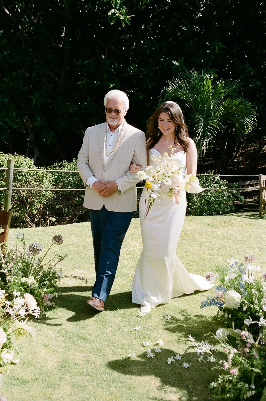 A bride, wearing a white wedding dress and holding a bouquet, walks arm-in-arm with an older man in a tan blazer and sunglasses during a wedding ceremony outdoors.