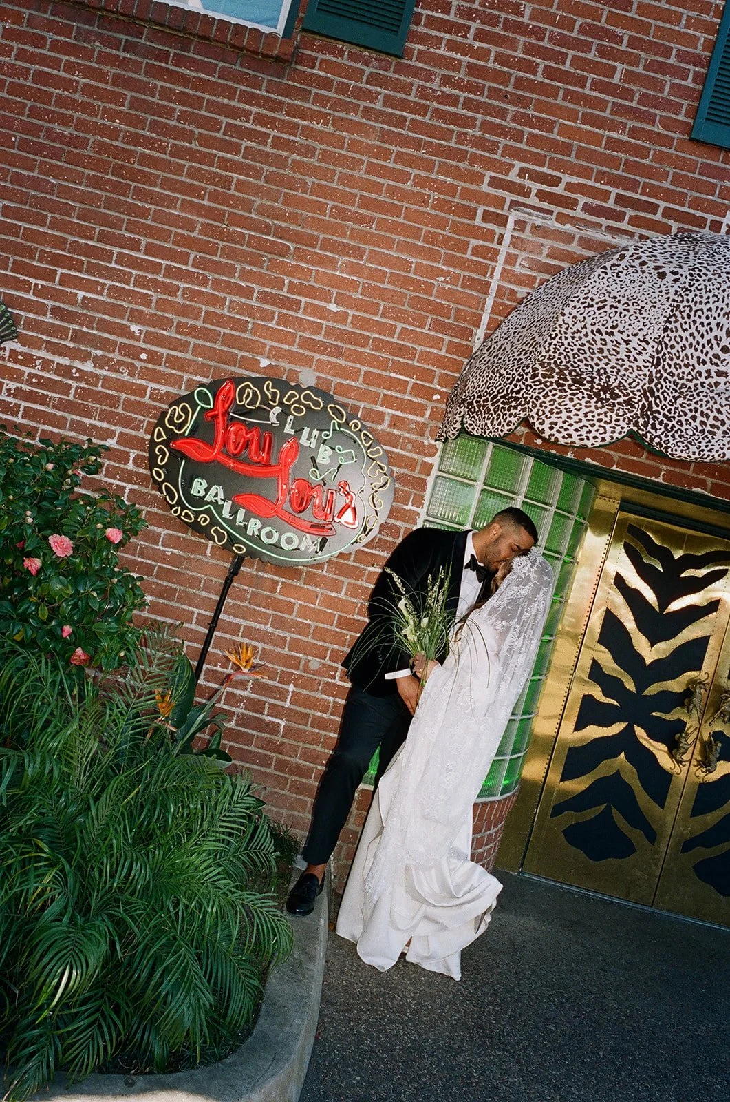 A couple dressed in formal wedding attire sharing a kiss outside a bar or lounge with a neon sign that reads "Love Love" and "Bar Lounge" in red and white. There is a leopard print umbrella, green plants, and a gold-colored door with black designs.