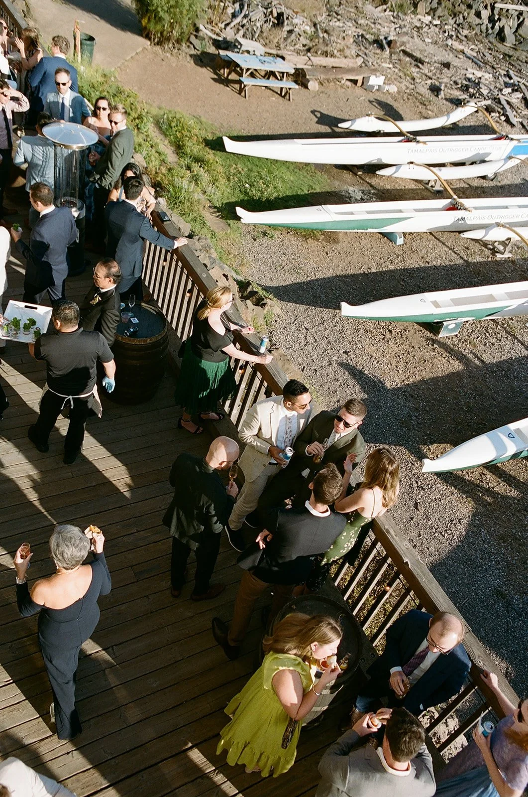 People socializing on a wooden deck at a gathering or party, with boats stored on land nearby.