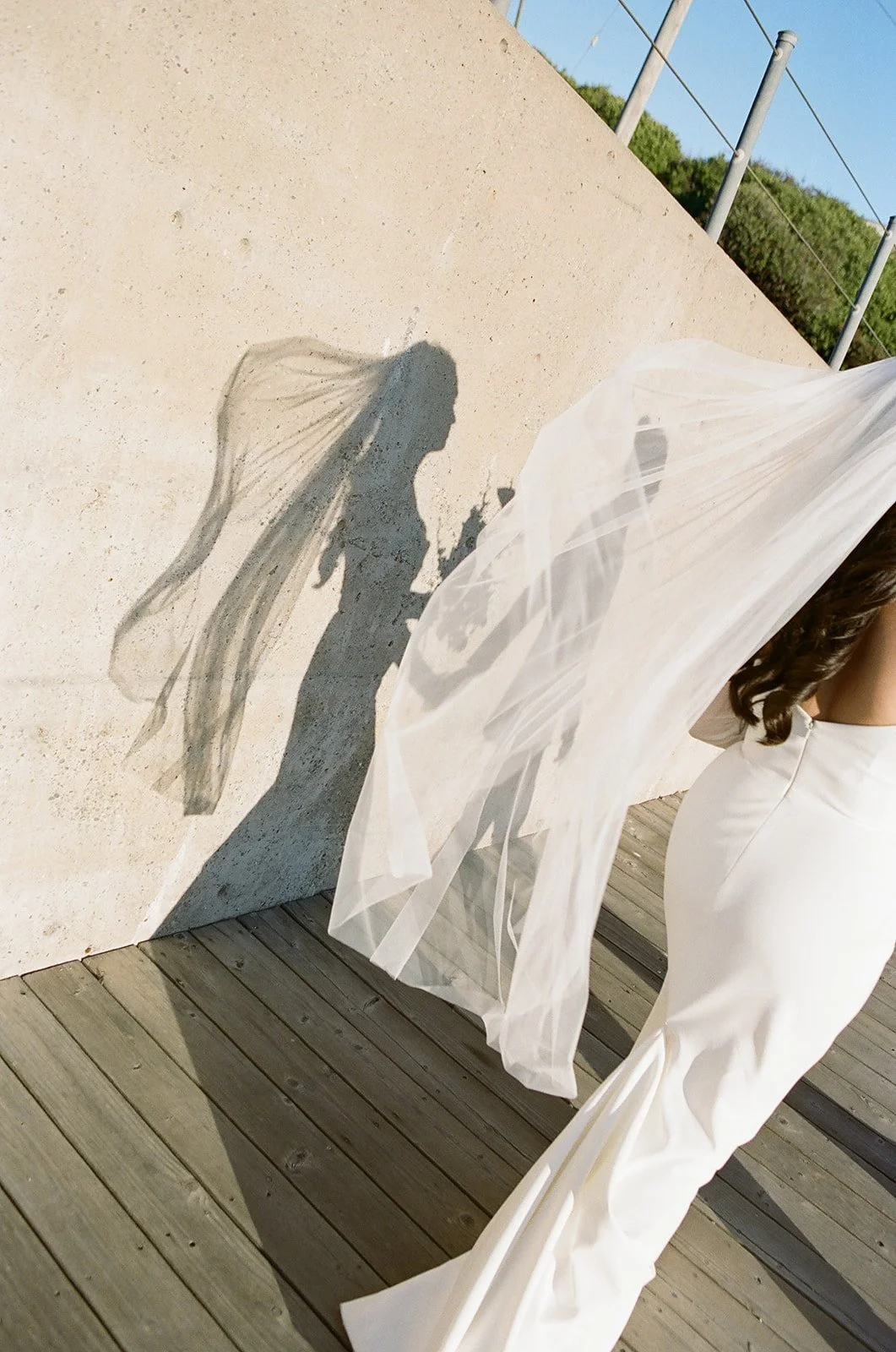 Shadow of a woman in a wedding dress with veil cast on a concrete wall, with woman partially visible in white dress on a wooden deck.