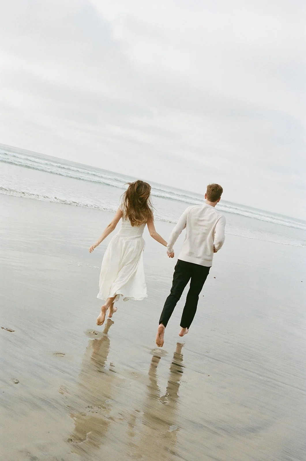 A couple holding hands and running on the beach with waves and cloudy sky in the background.