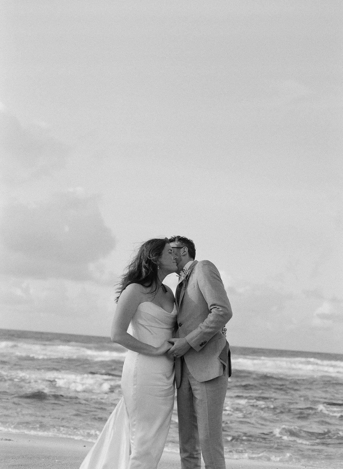 A couple in wedding attire sharing a kiss on the beach with ocean waves and cloudy sky in the background.