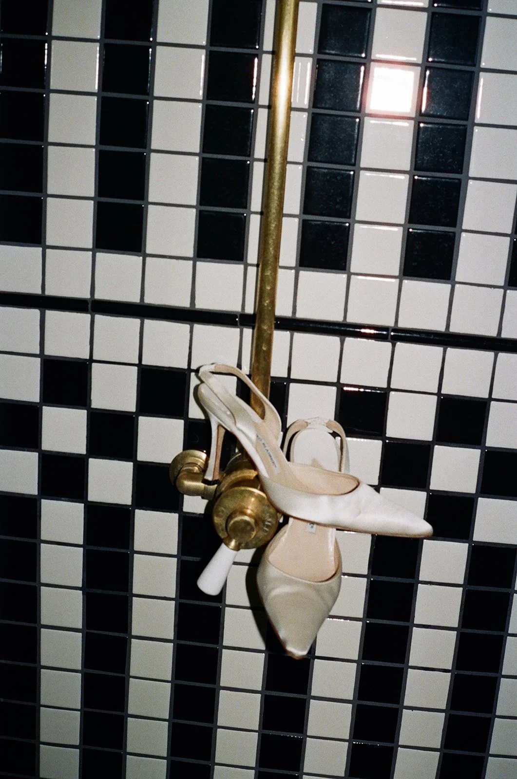 A pair of white high-heeled shoes hanging from a brass pipe against a black and white tiled wall.