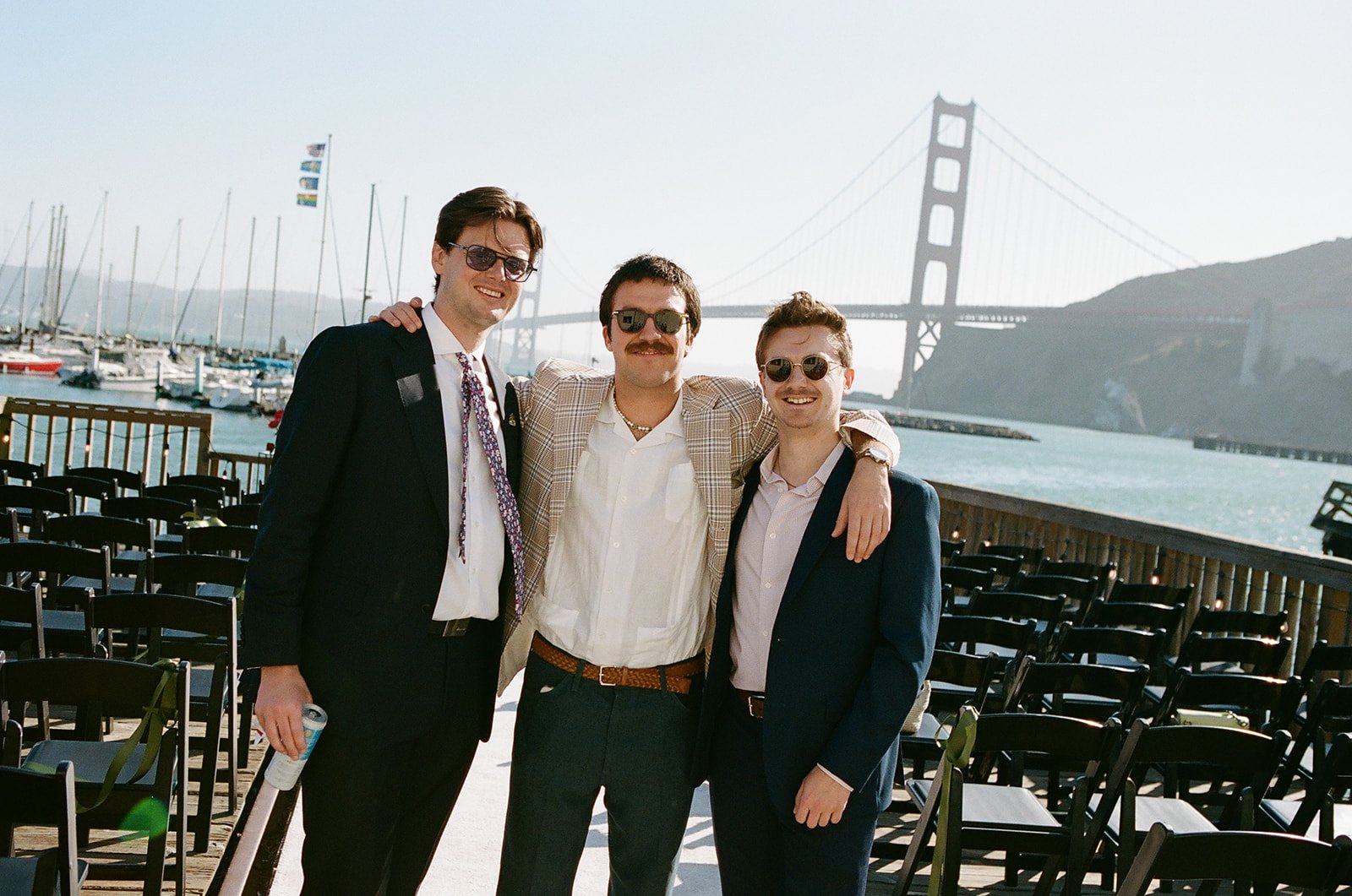 Three men in suits with sunglasses smiling, standing close together with arms around each other, in front of a view of the Golden Gate Bridge and boats docked at a marina.
