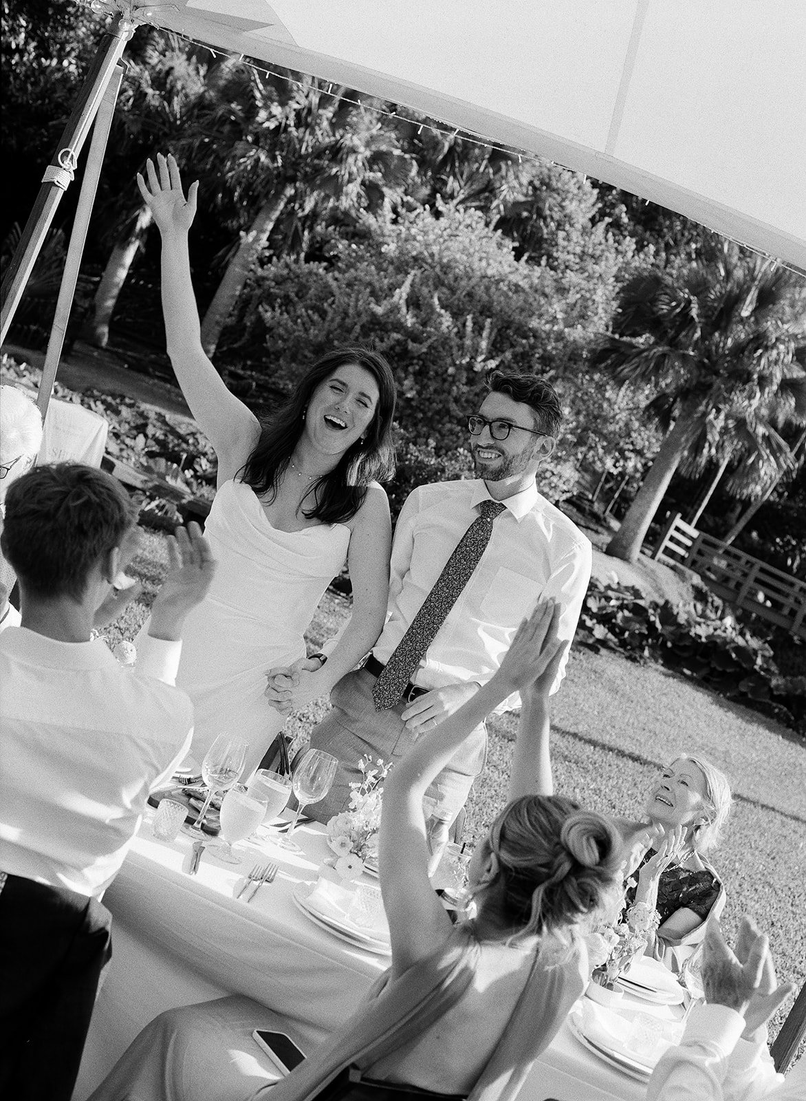 Black and white photo of a joyous outdoor celebration with a woman in a white dress and a man in glasses and a tie smiling, and others around them clapping and celebrating. Trees and outdoor decor are visible in the background.