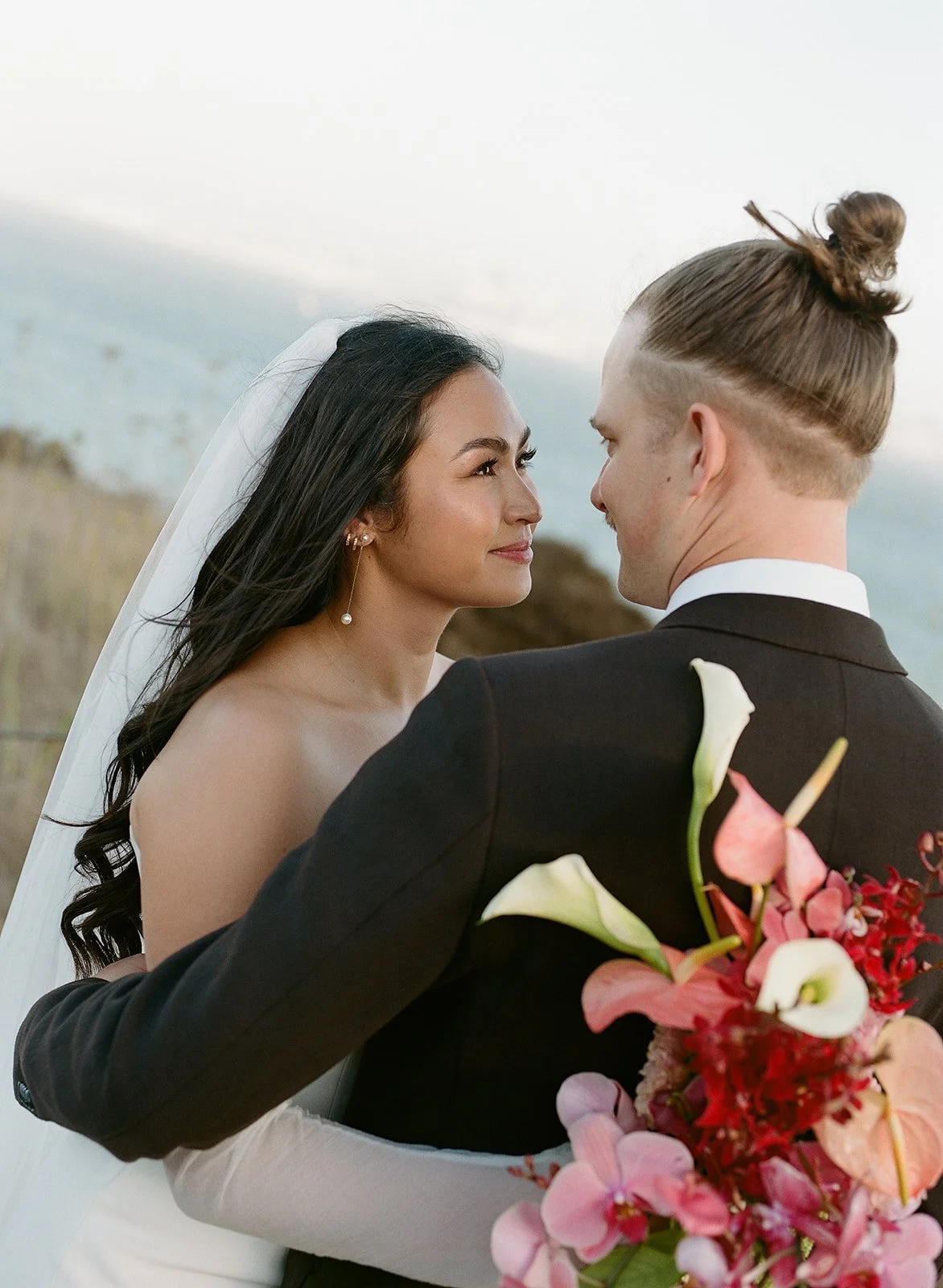 A bride and groom face each other closely outdoors, with the bride holding a bouquet of pink and white flowers, over a natural landscape background.