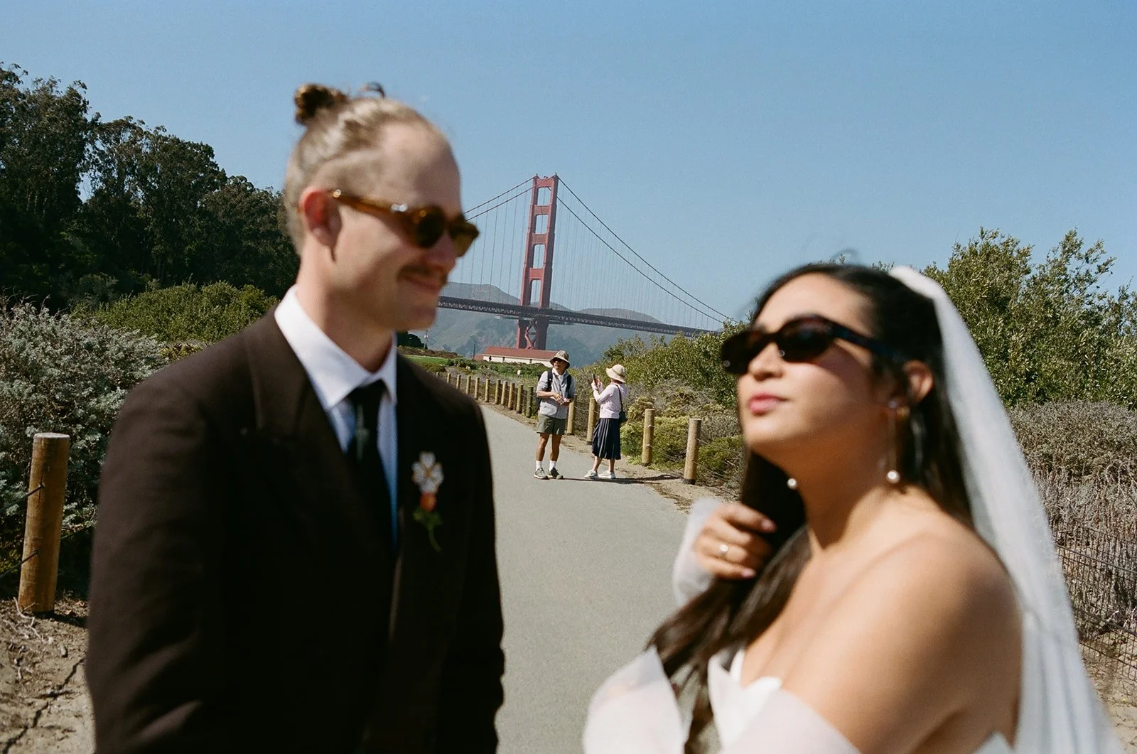 A couple is posing in front of the Golden Gate Bridge in San Francisco on a sunny day, with two tourists taking photos in the background.