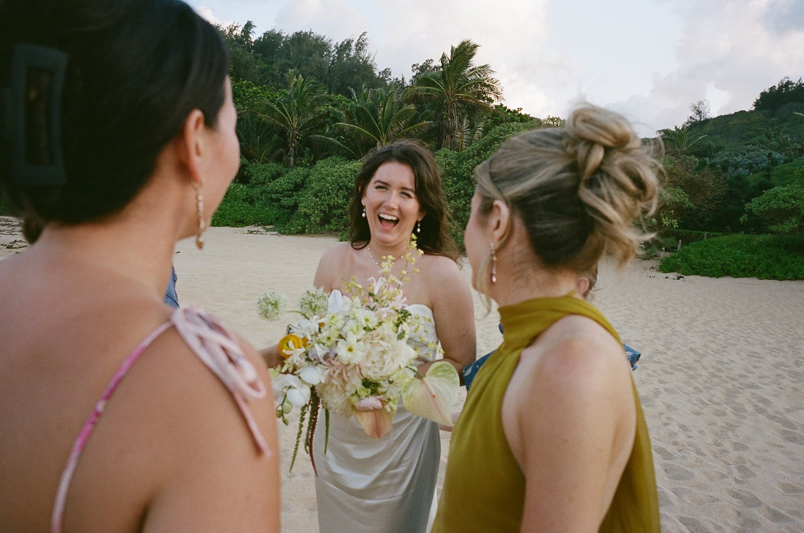 Group of women celebrating on a sandy beach with lush green trees and cloudy sky in the background.