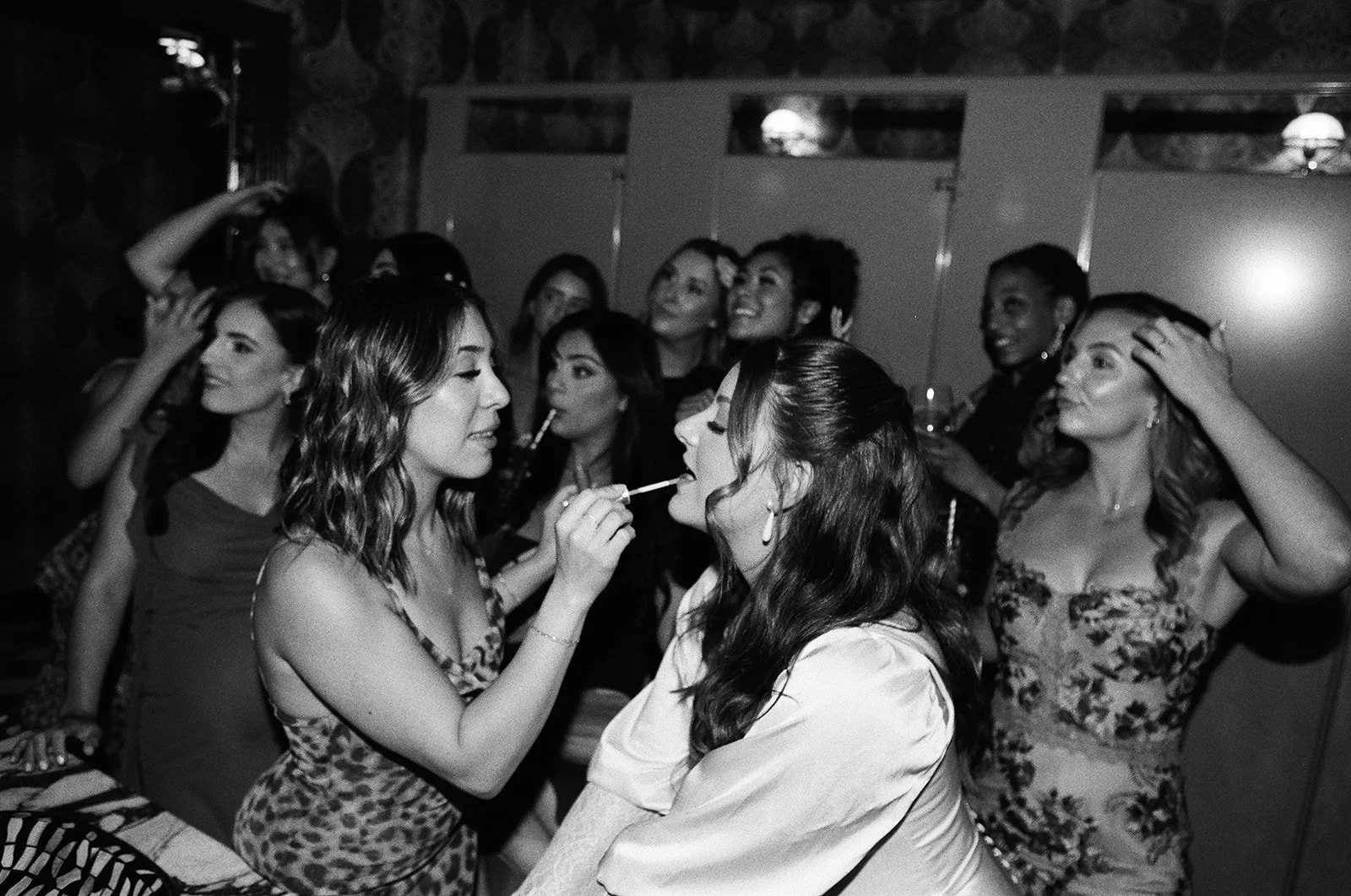 A group of women at a party, with one woman applying lipstick to another, while others are smiling and interacting in the background.