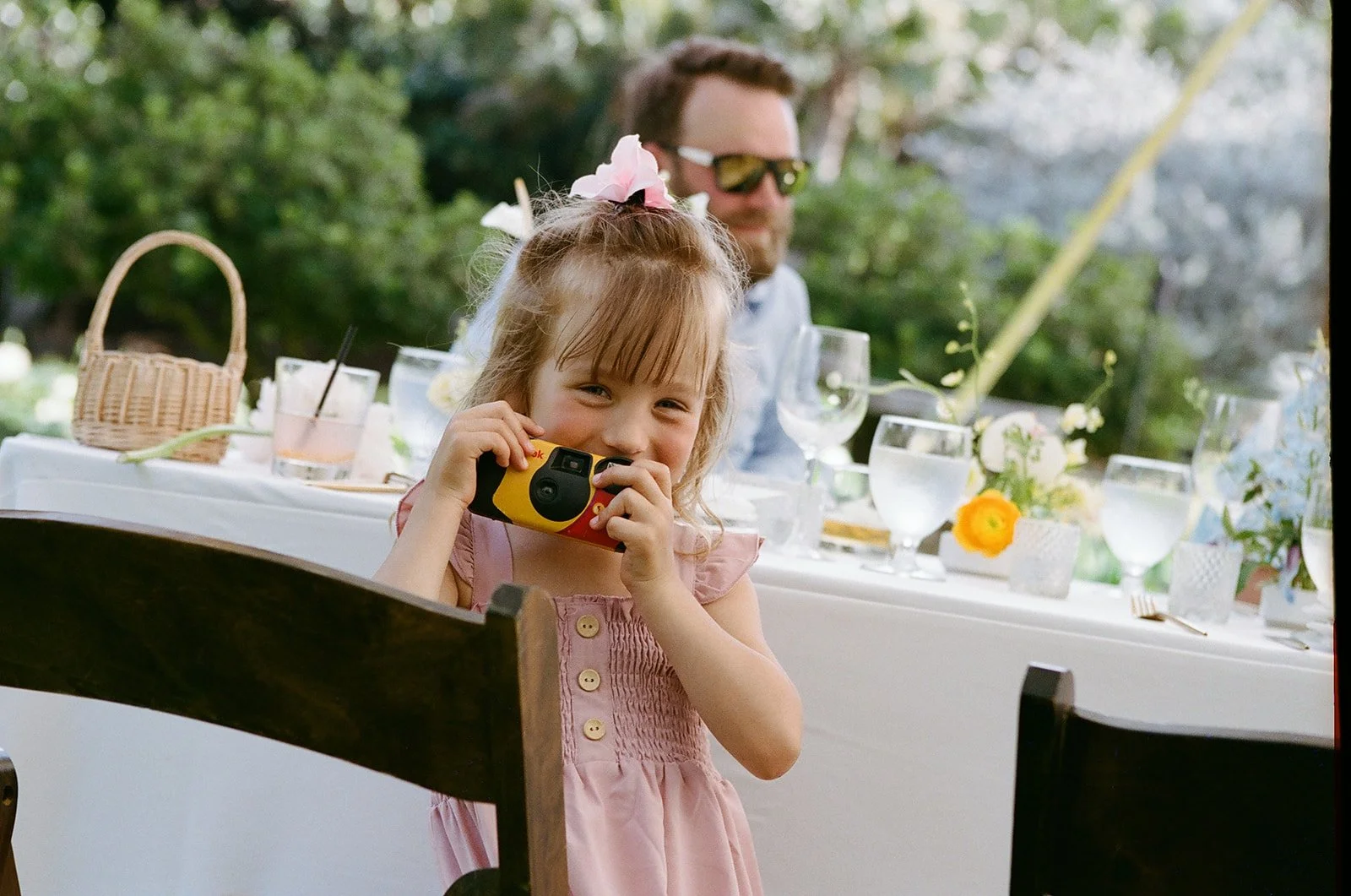 A young girl with light brown hair, wearing a pink dress with buttons, is smiling and holding a yellow and red toy camera. She has pink and white flowers in her hair. Behind her, a man with sunglasses and light-colored shirt is sitting at a table out