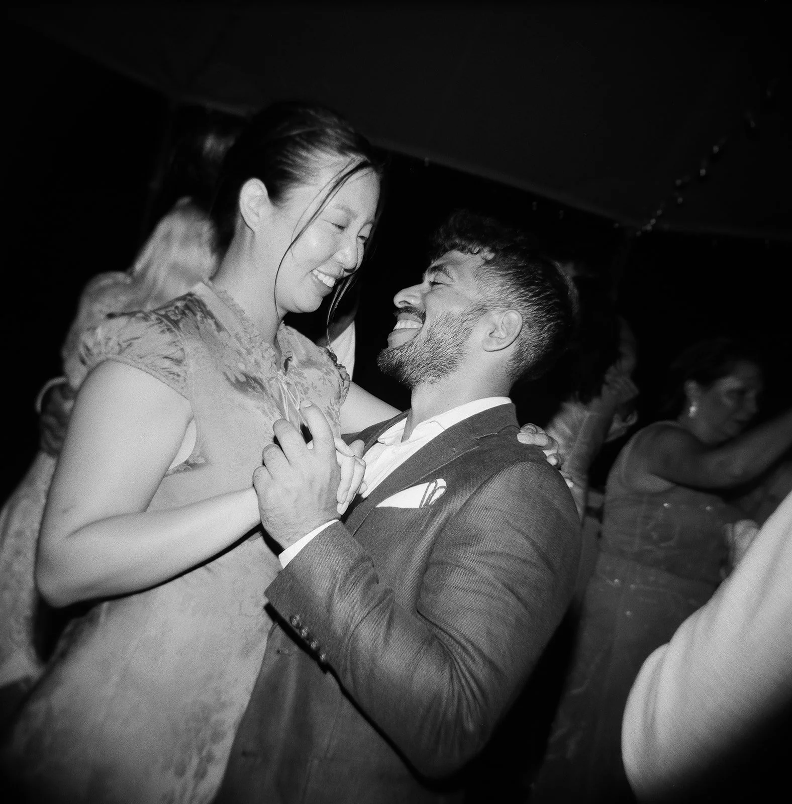 A man and woman dance closely, smiling, at a party or celebration, surrounded by other people in a dark room.