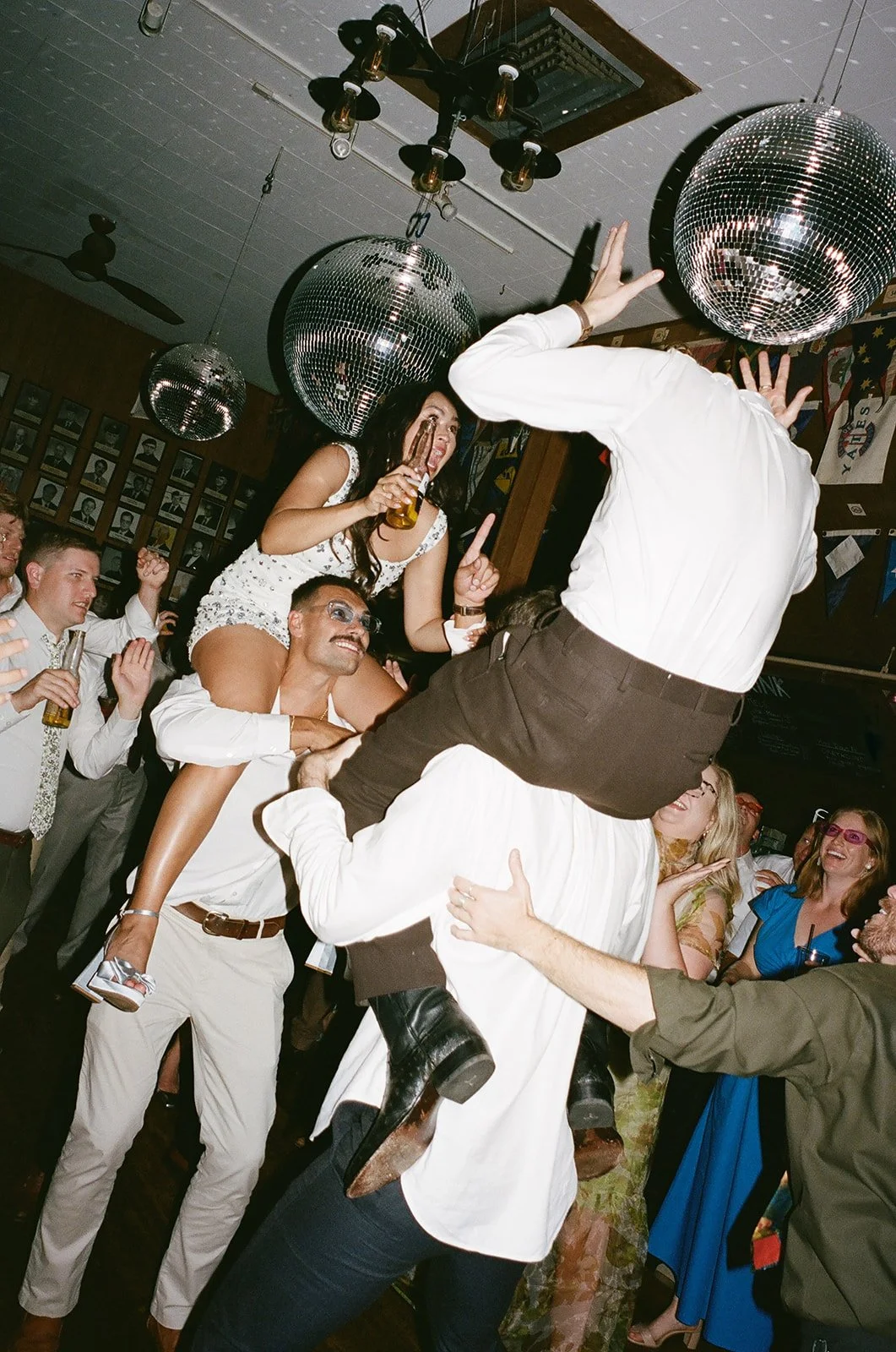 People dancing and celebrating at a lively party, with a woman sitting on a man's shoulders, holding a drink, and others dancing around under disco balls.