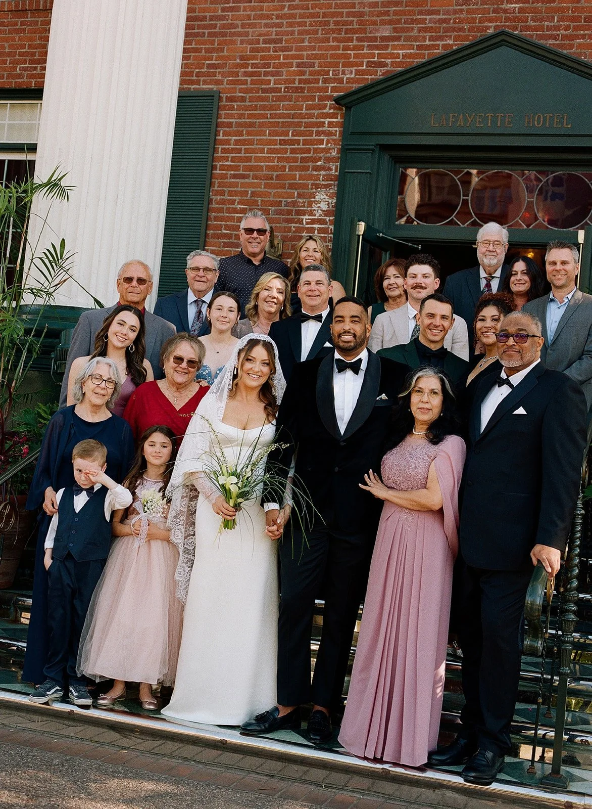 A wedding celebration with the bride and groom standing in front, surrounded by family and friends outside the Lafayette Hotel.