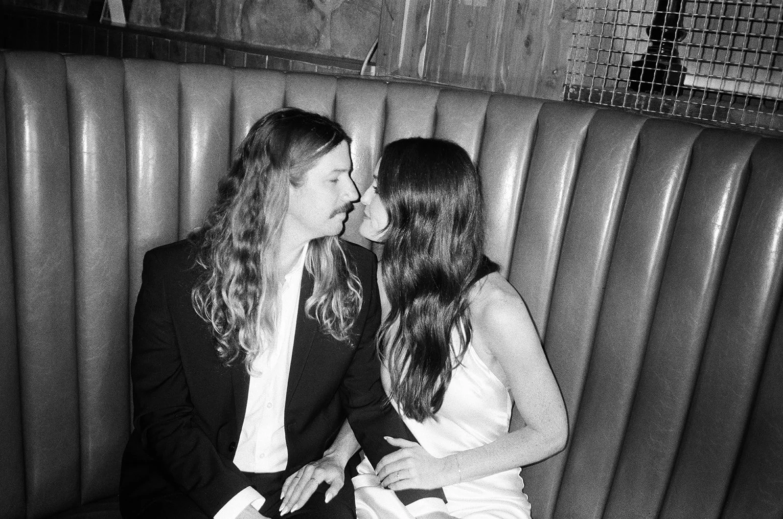 A man with long hair and a mustache and a woman with long wavy hair are sitting close together on a bench in a dimly lit room, about to kiss.
