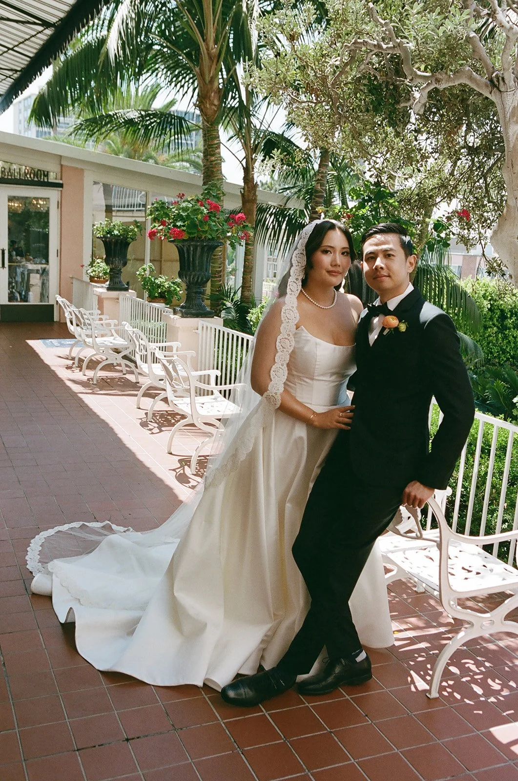 A bride and groom posing outdoors on a sunny day, with lush greenery and potted plants in the background. The bride is wearing a white wedding gown with lace detailing and a veil, and the groom is dressed in a black tuxedo with a bow tie and boutonni