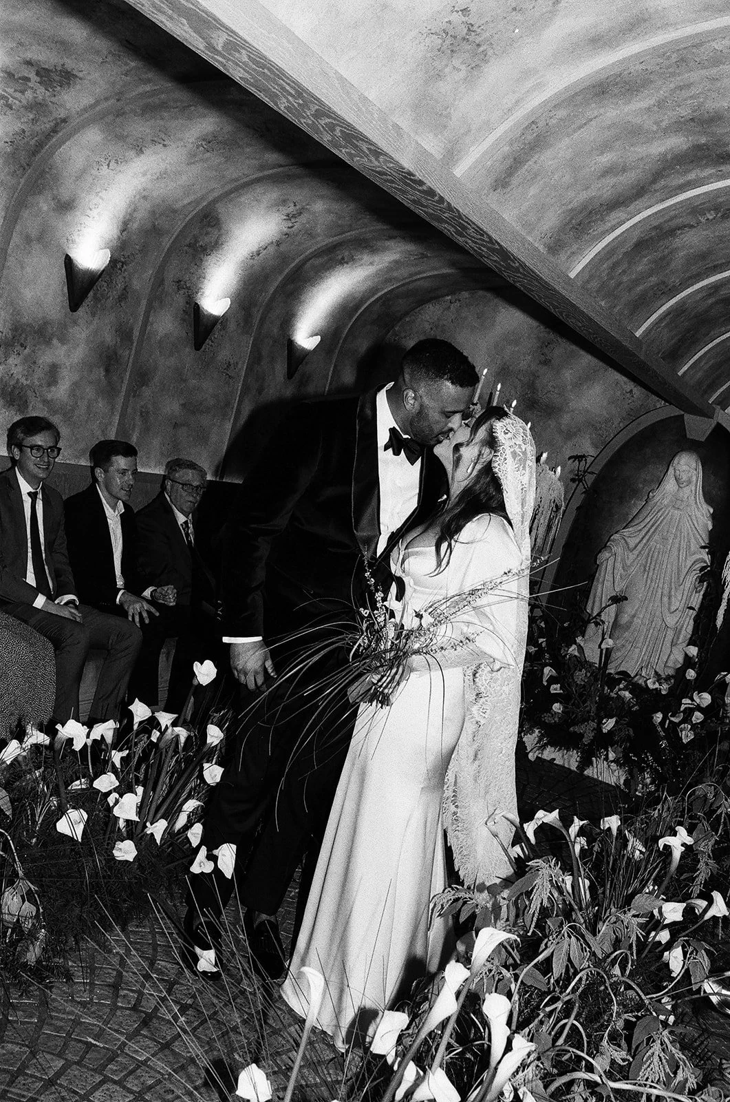 A black-and-white photo of a wedding ceremony showing a bride and groom kissing, with guests seated in the background inside a church or chapel.