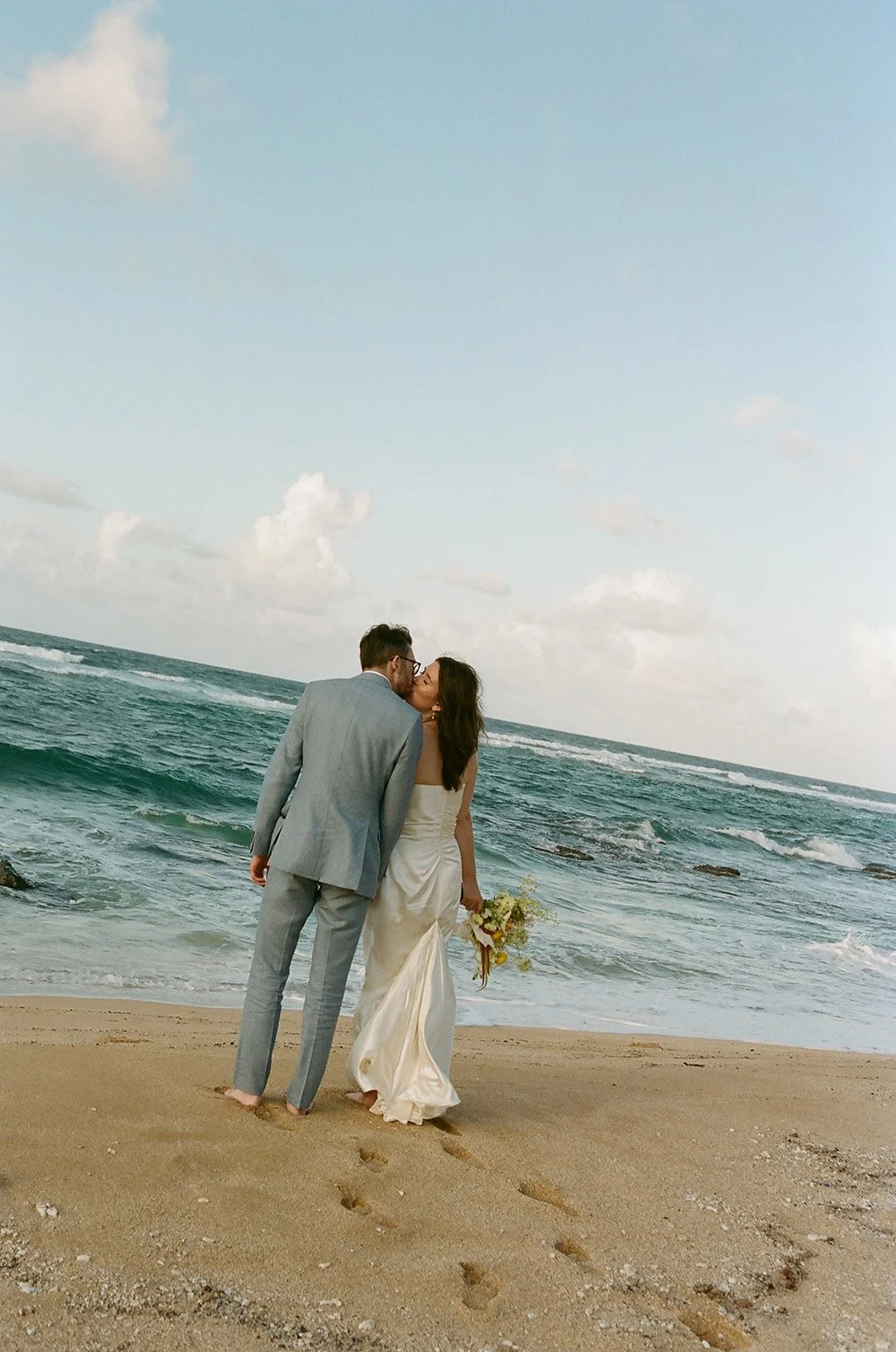 A couple in wedding attire share a kiss on a sandy beach with ocean waves in the background.