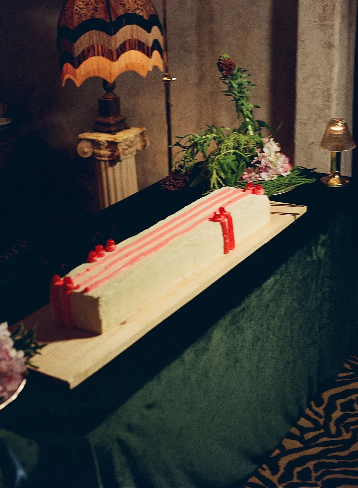A rectangular birthday cake with white and pink icing, decorated with red candies and a red number 1 candle on top, placed on a wooden board on a table with a black and green tablecloth, surrounded by flowers and small lamps.
