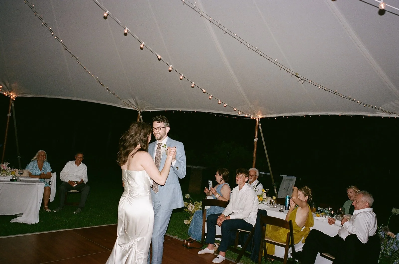 A couple dancing under a white canopy at a wedding reception at night, with guests sitting at tables watching.