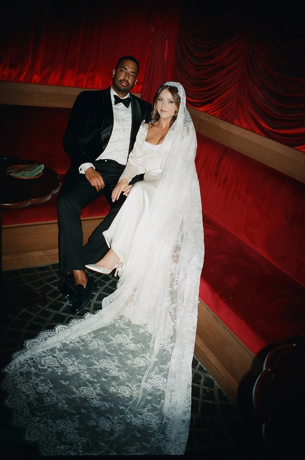 A groom in a tuxedo sitting next to a bride in a white wedding dress with a long lace veil, seated on a red velvet bench in a dimly lit room with red curtains.