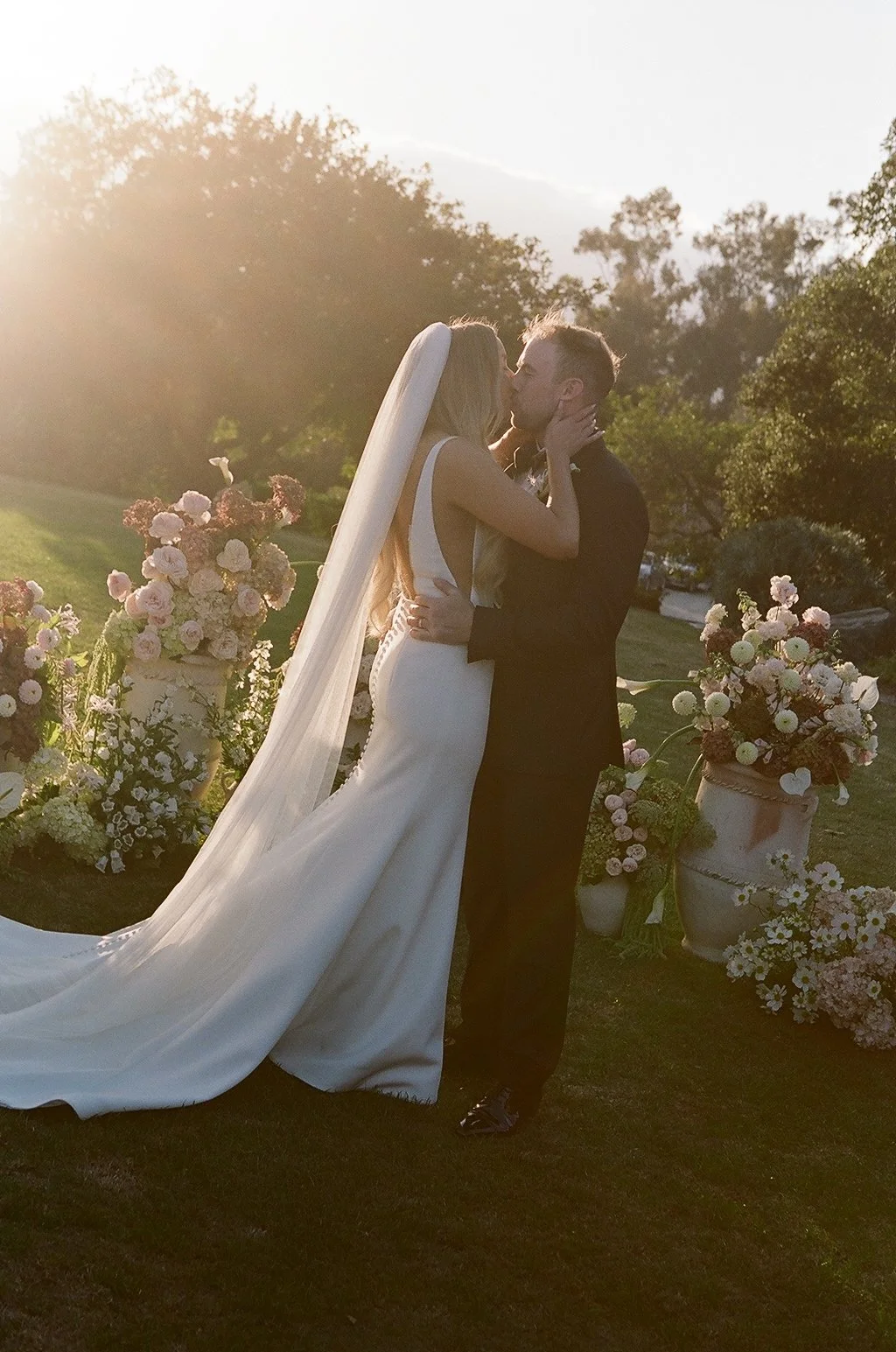 A bride and groom sharing a kiss during their wedding ceremony outdoors at sunset, with floral arrangements in large urns around them.