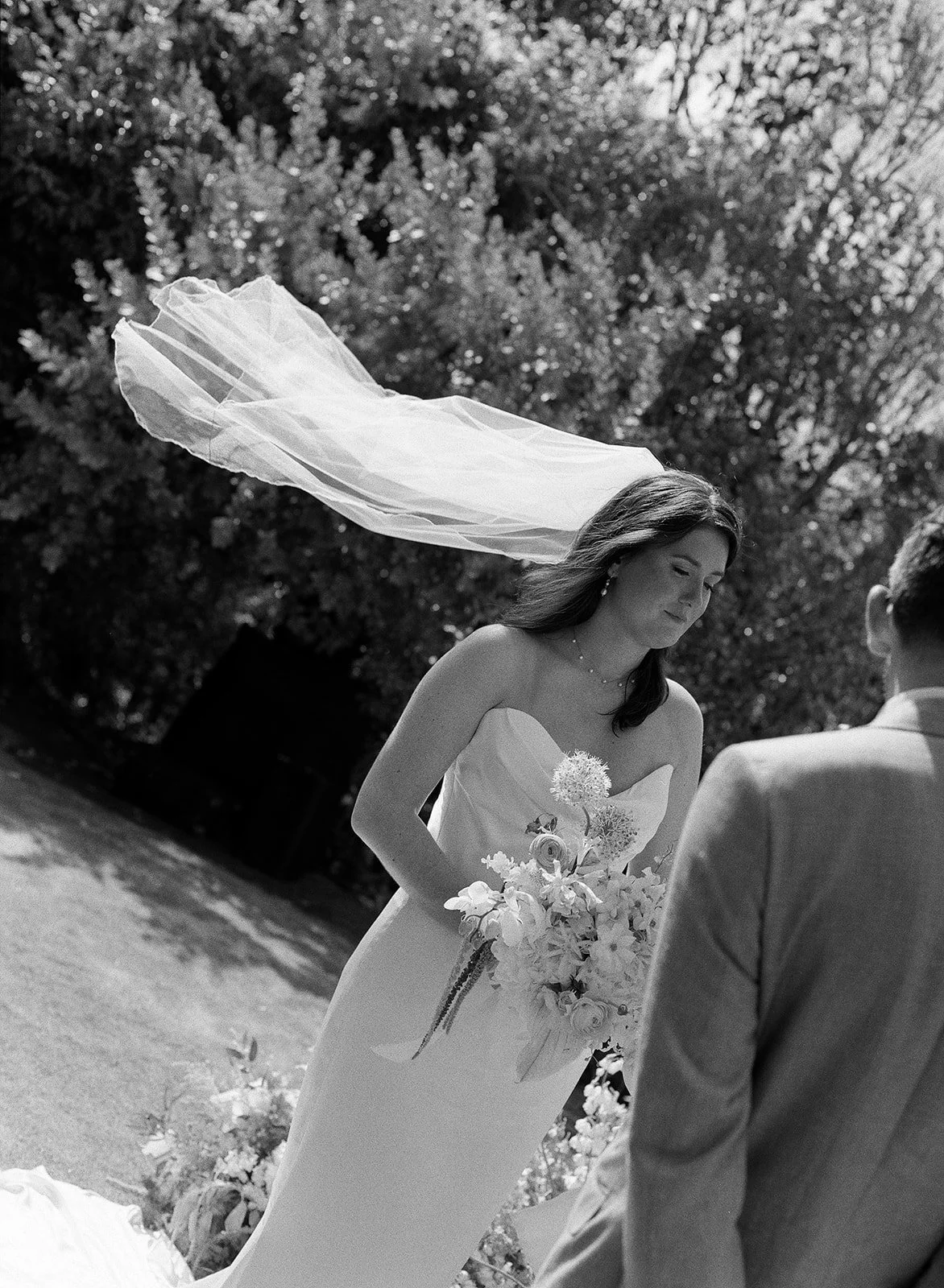 Black and white photo of a bride in a strapless wedding dress with her eyes closed, holding a bouquet of flowers, outdoors with trees in the background, and a veil blowing in the wind.