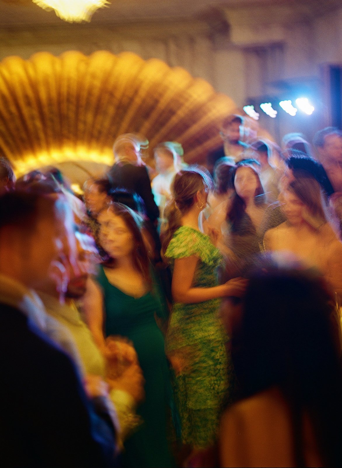 People dancing and socializing at a party or event in a dimly lit room with colorful lighting and a curved wooden ceiling.