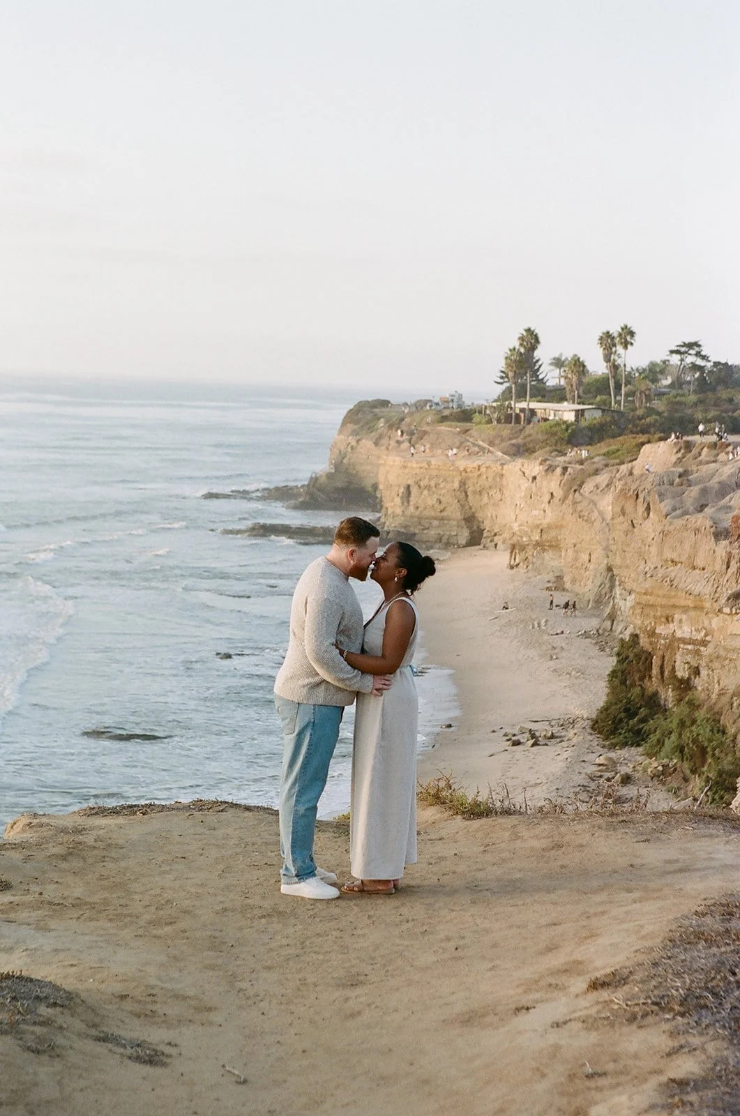 A couple embracing and about to kiss on a cliff overlooking the beach with ocean waves and rocky cliffs, with palm trees and a few people in the background.