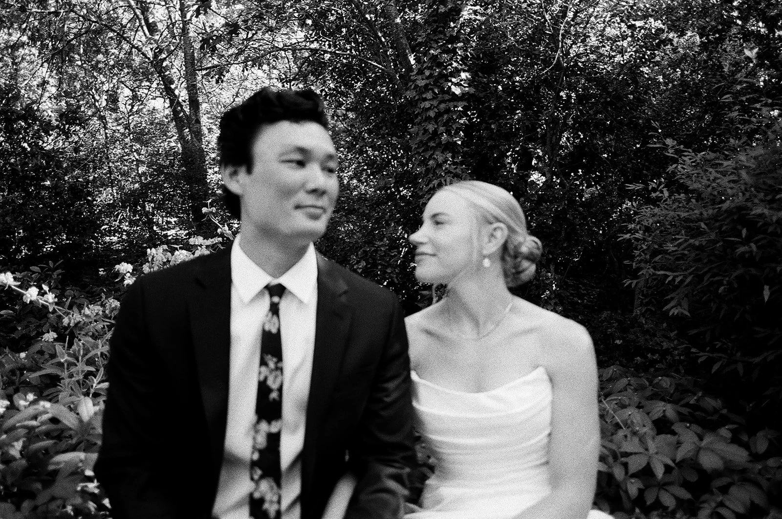 A black-and-white photo of a couple in wedding attire outdoors, with trees and bushes in the background. The man is wearing a suit and tie, and the woman is wearing a strapless dress with her hair styled in an updo. She is looking at him with a smile