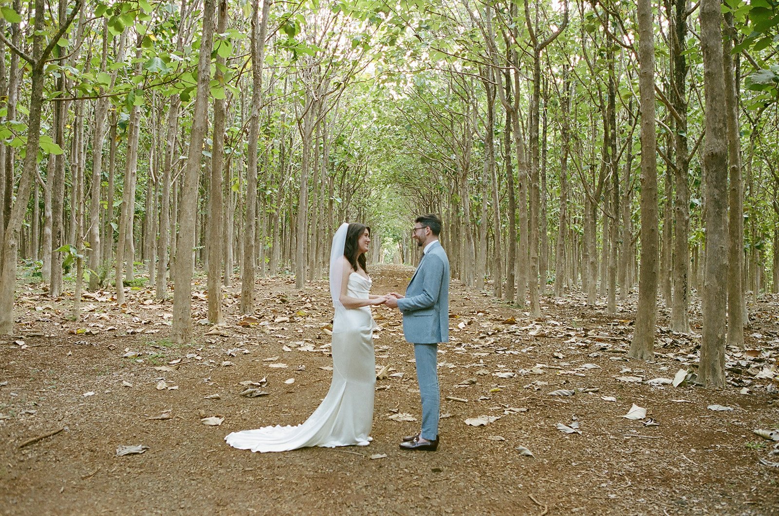 A bride and groom holding hands and facing each other in a forest with tall trees and fallen leaves.