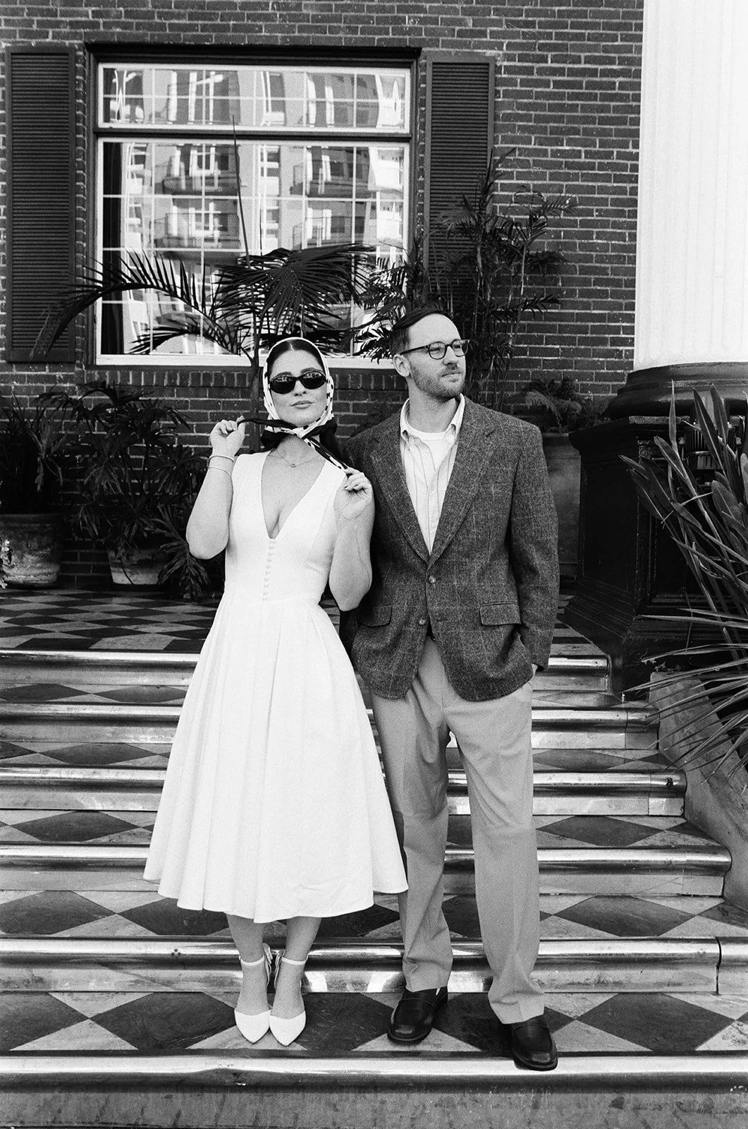A woman in a vintage white dress with a deep neckline and a man in a blazer and slacks standing on steps outside a brick building with plants and a window.