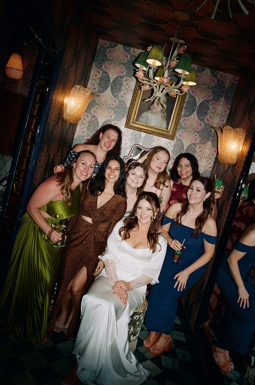 A group of women dressed in elegant evening wear posing together in a decorated room with ornate wallpaper, chandelier, and wall sconces.