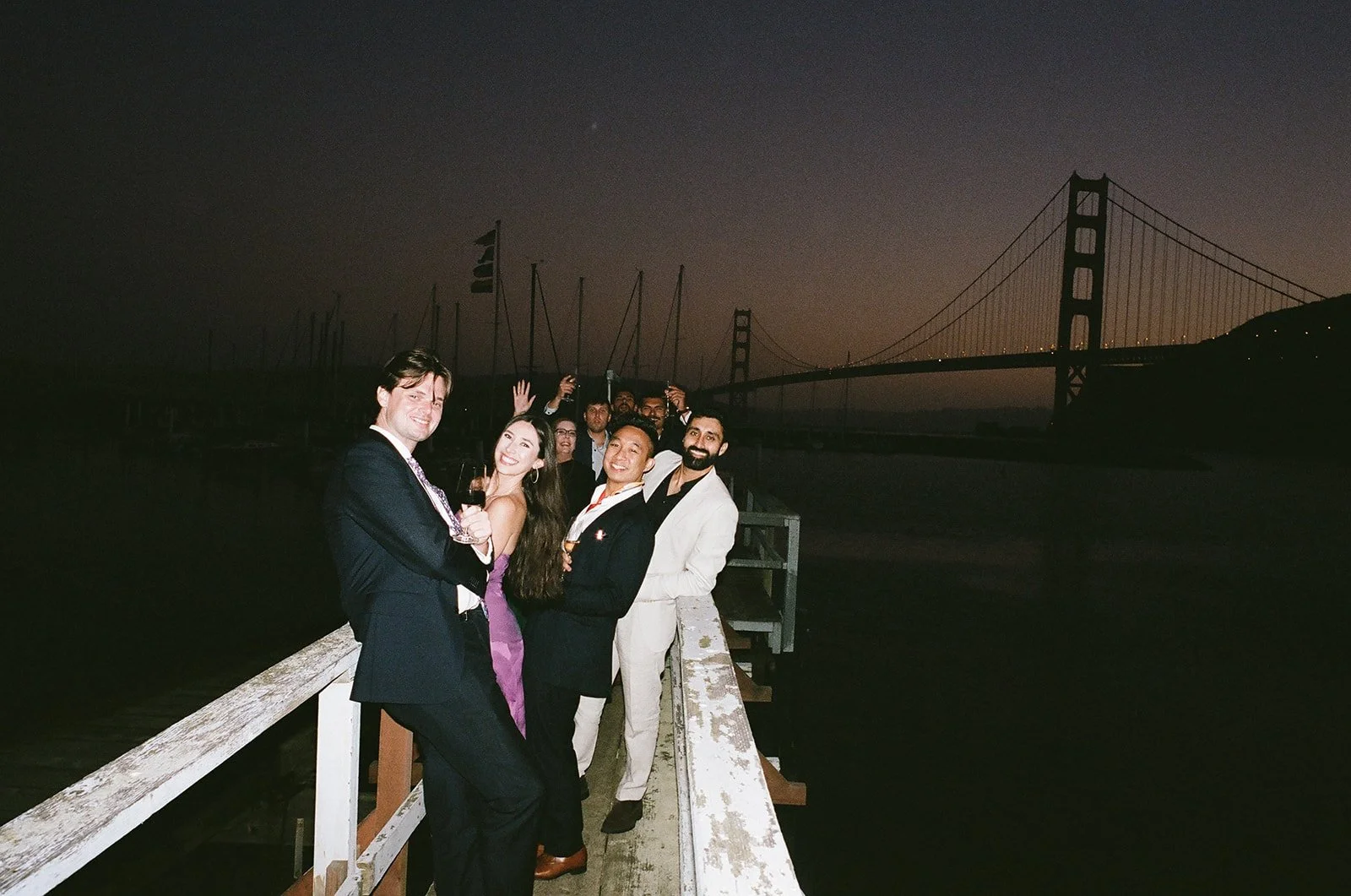 Group of friends in formal attire on a pier at night, with the Golden Gate Bridge in the background.