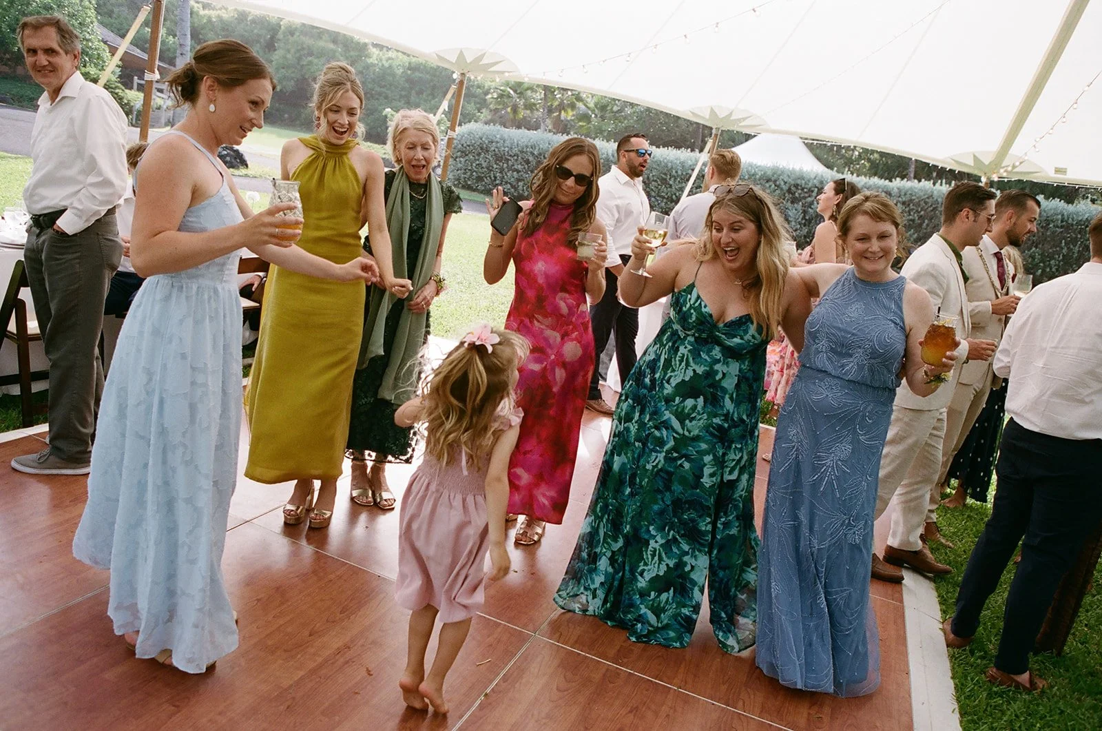 People dancing and celebrating at an outdoor wedding reception under a tent, wearing colorful dresses and holding drinks.