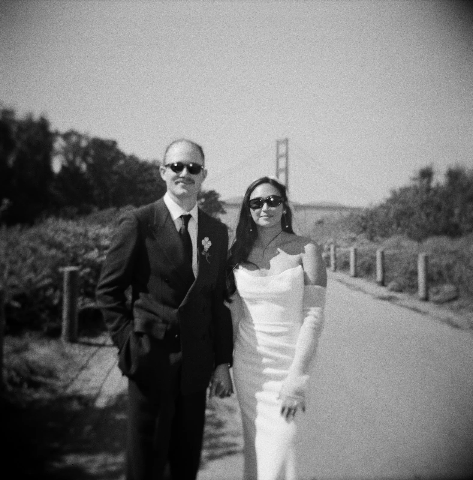 A black and white photo of a man and woman standing outdoors on a pathway, with the Golden Gate Bridge in the background. They are wearing sunglasses and formal attire, with the man in a suit and the woman in a white dress.
