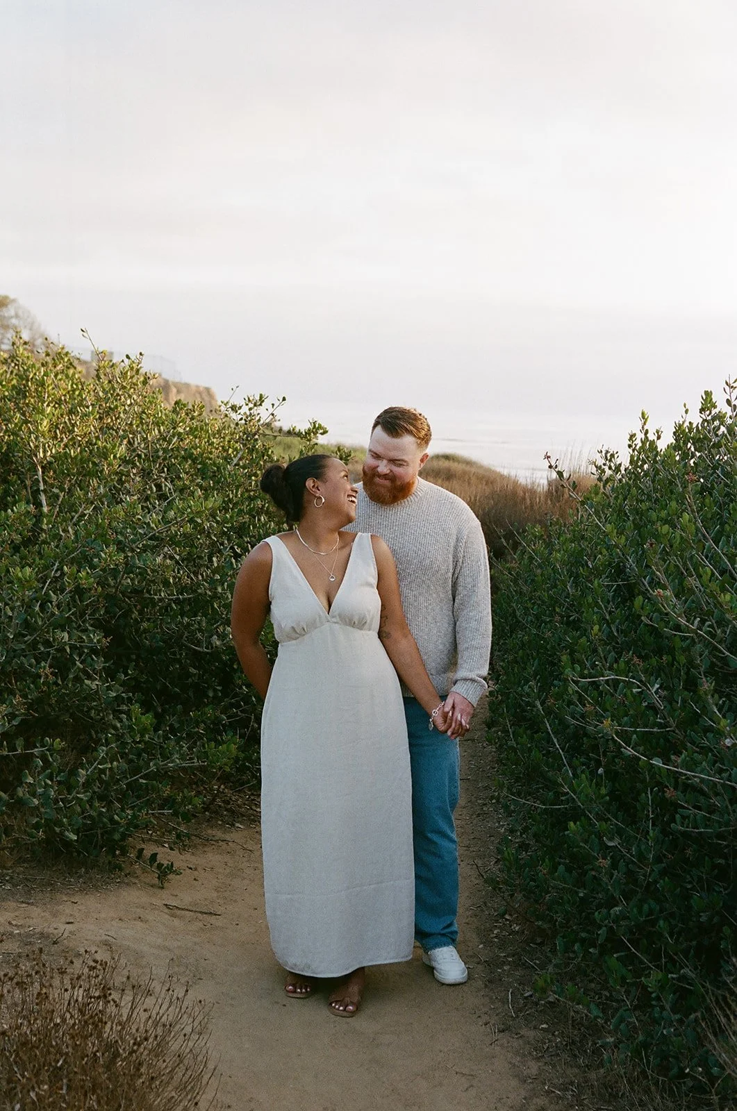 A happy couple holding hands and smiling at each other during a walk through a nature trail with bushes and trees, with the ocean in the background.