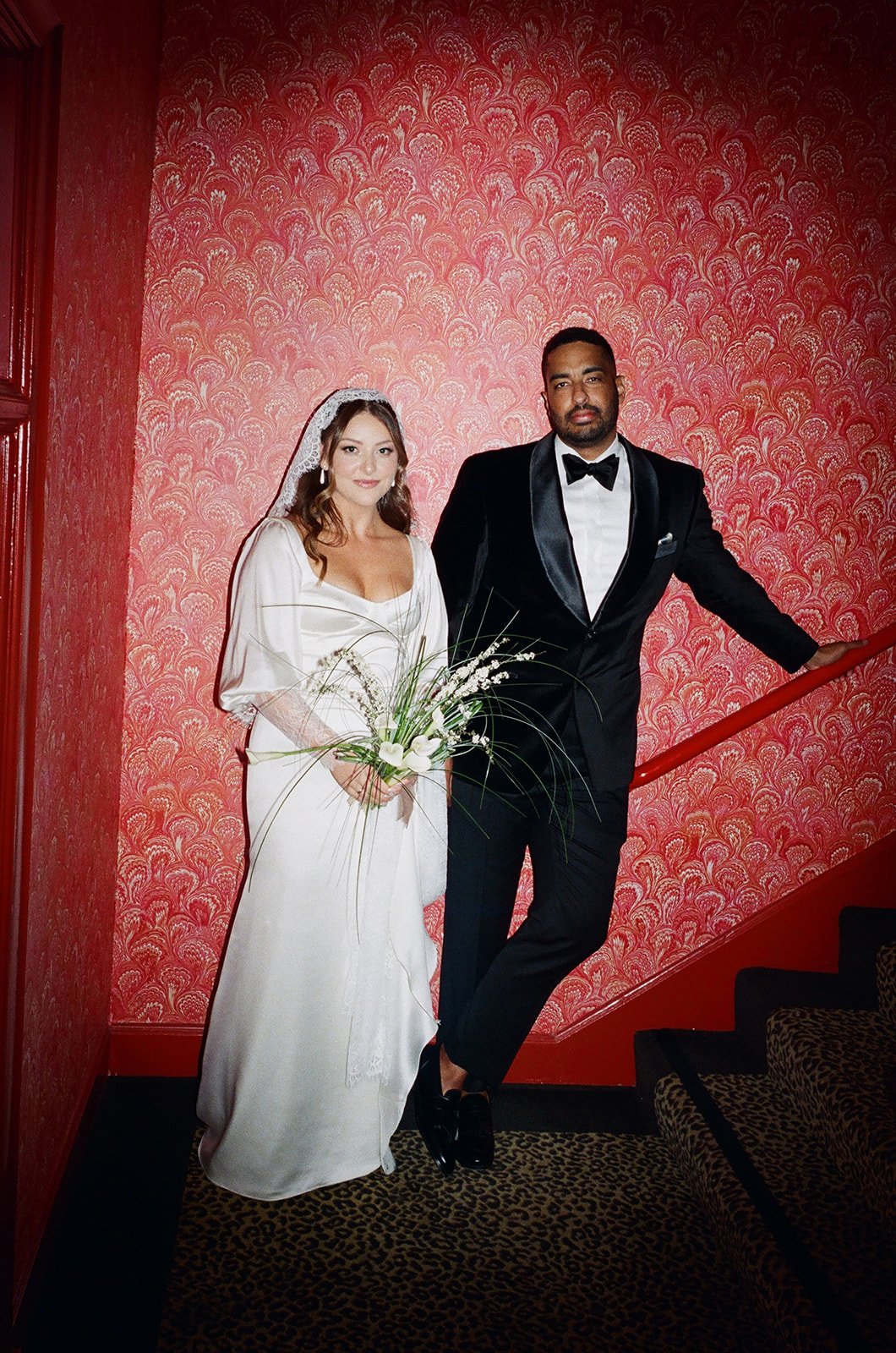 A bride and groom posing on a staircase with a red patterned wallpaper background. The bride is holding a bouquet and wearing a white wedding dress with a veil, while the groom is in a black tuxedo with a bow tie.