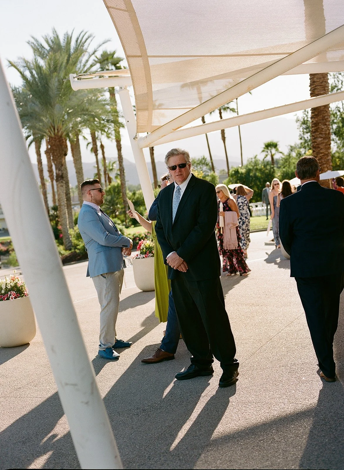 People dressed in formal attire standing outdoors near palm trees under a shaded area at a sunny event.