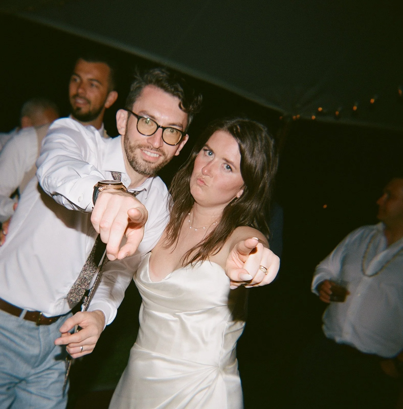A man and woman pointing at the camera at a social event, with other people in the background.