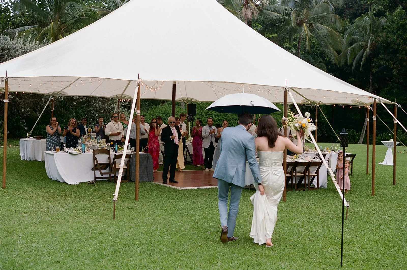 A couple walks towards a wedding reception under a large white tent on a grassy field. The bride holds a bouquet of flowers and is dressed in a white gown, while the groom is in a light blue suit holding an umbrella. Guests stand inside the tent, cla